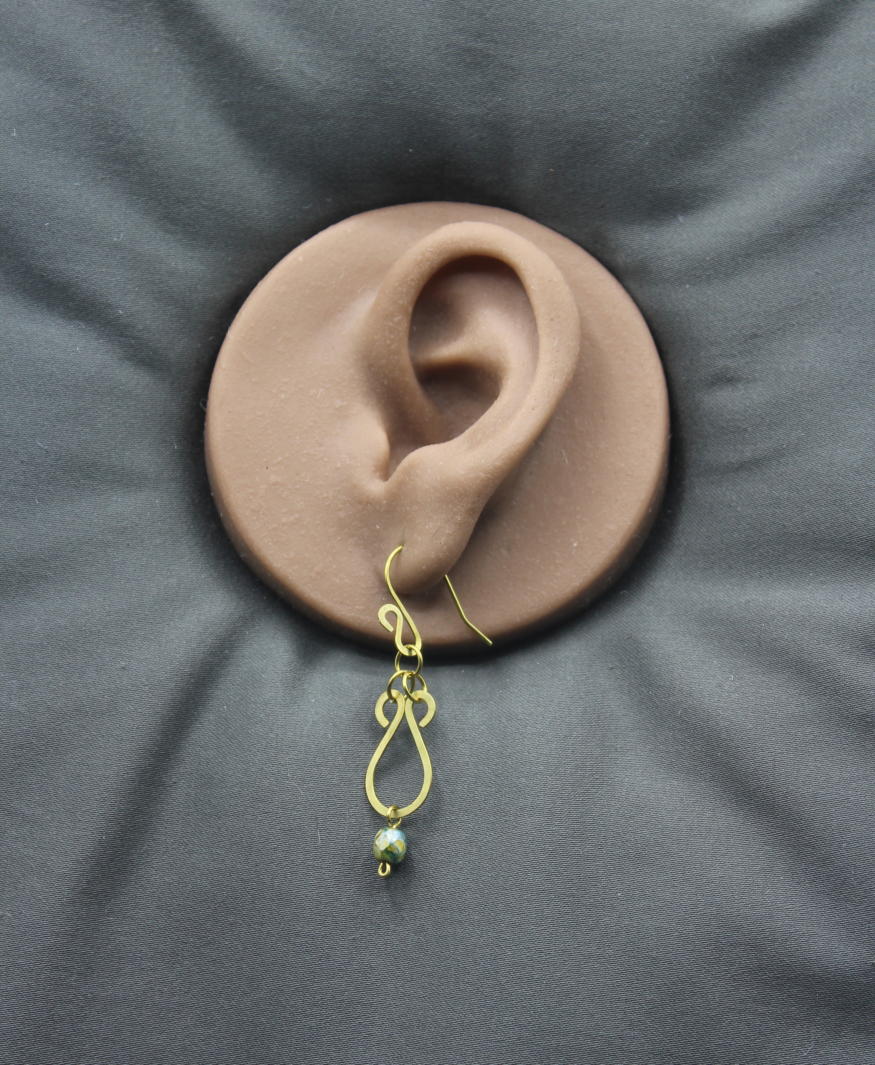 Hammered brass teardrop earrings with mottled yellow/blue bead drops on a clay model of an ear against a gray background
