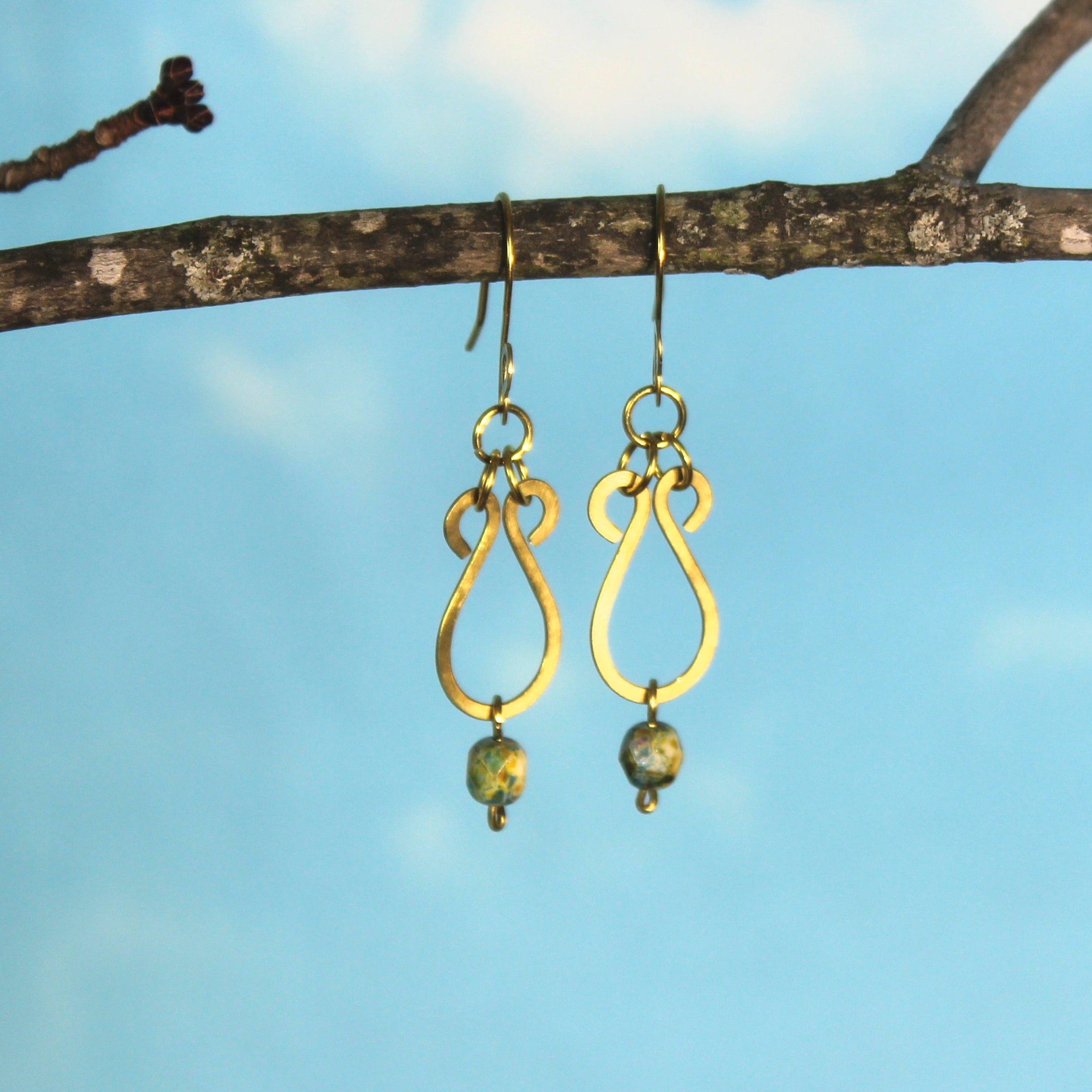Hammered brass teardrop earrings with mottled yellow/blue bead drops hanging from a branch against a blue sky.