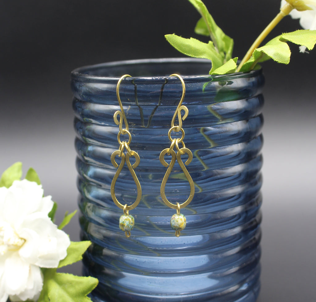 Hammered brass teardrop earrings with mottled yellow/blue bead drops on a blue glass vase with white flowers and green leaves.