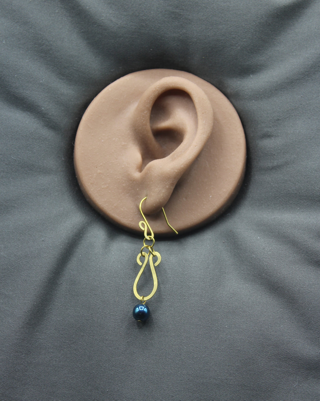 Hammered brass teardrop earring with a metallic dark teal bead drop on a clay ear model against a gray background