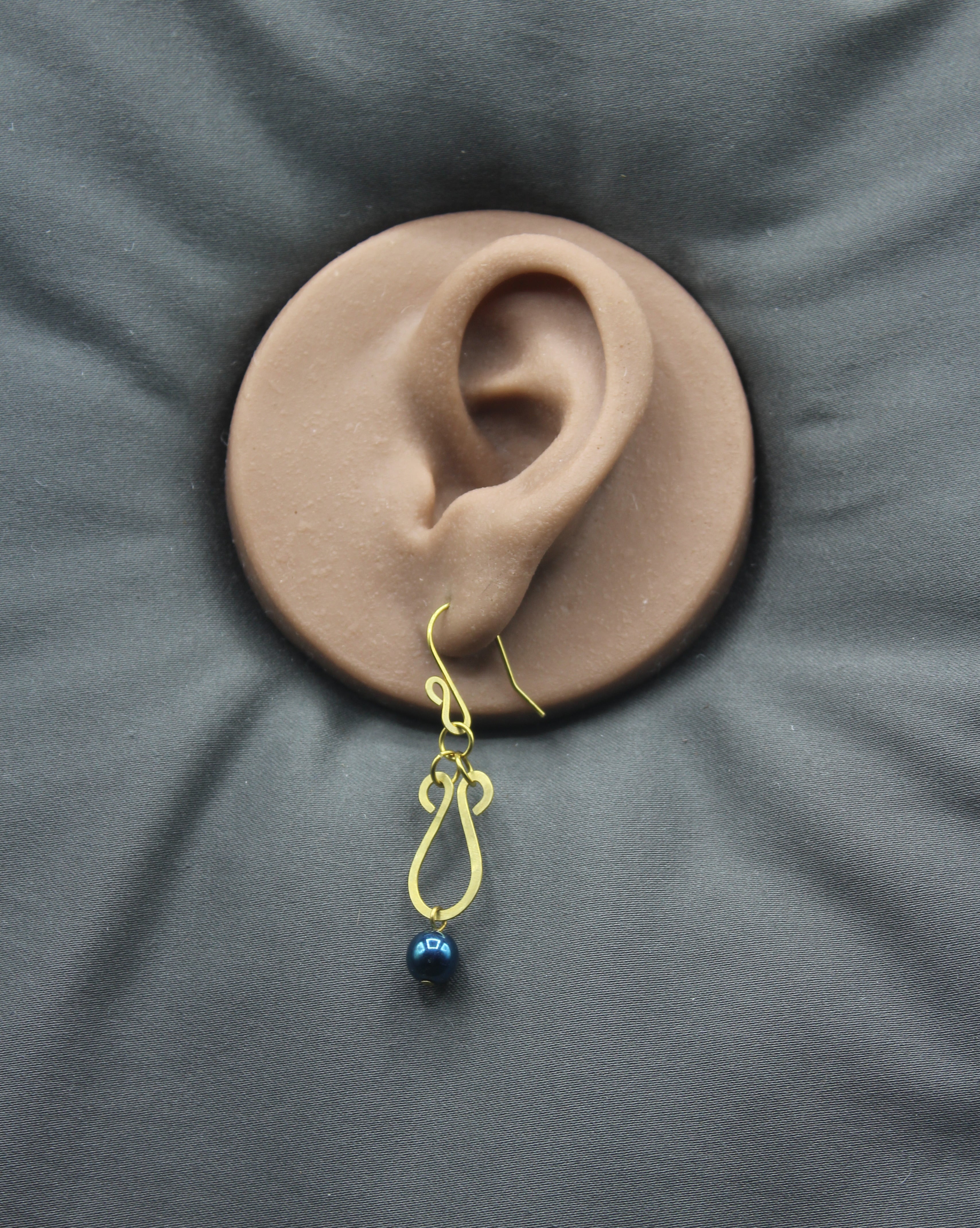 Hammered brass teardrop earring with a metallic dark teal bead drop on a clay ear model against a gray background