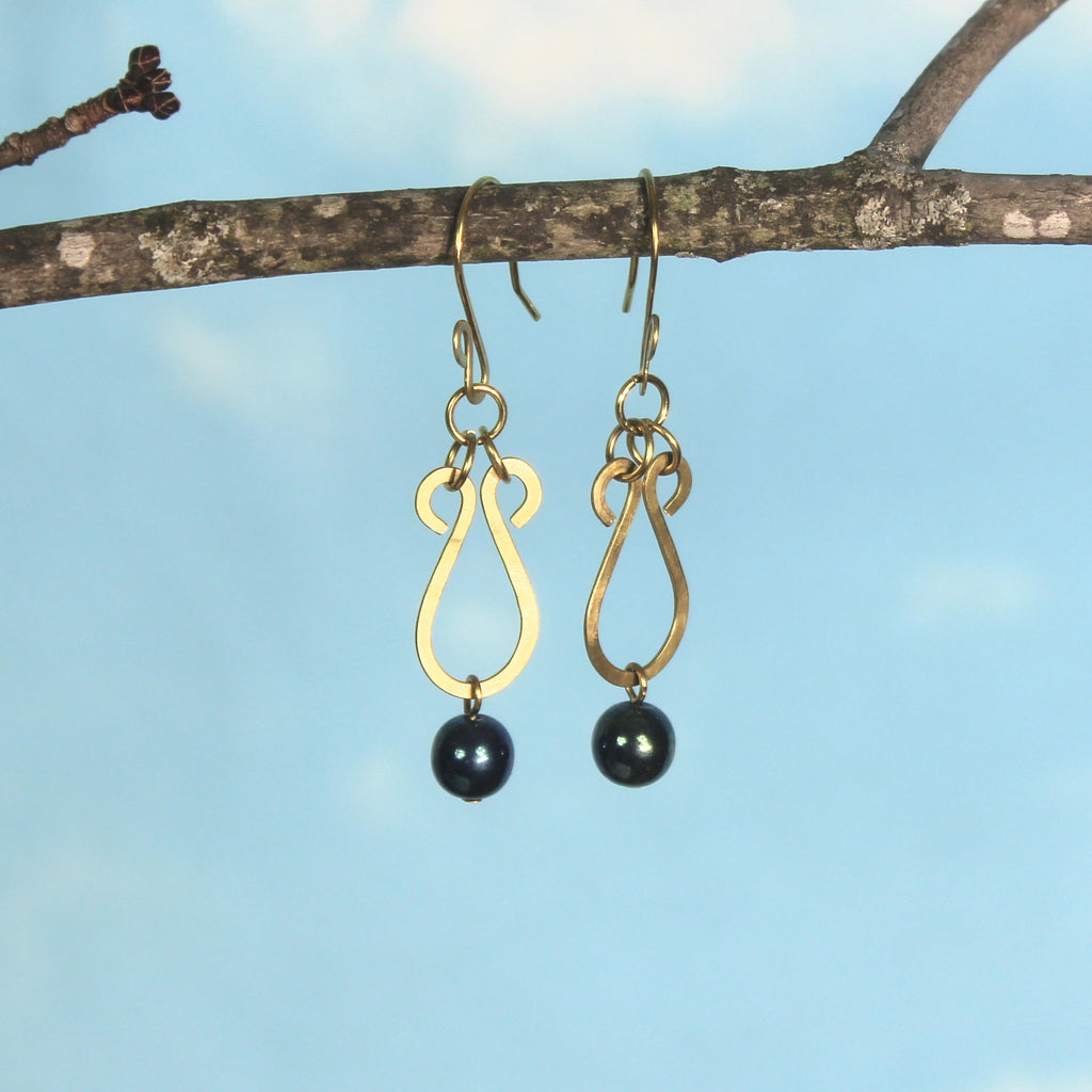 Hammered brass teardrop earrings with metallic dark teal bead drops hanging from a branch against a blue sky.