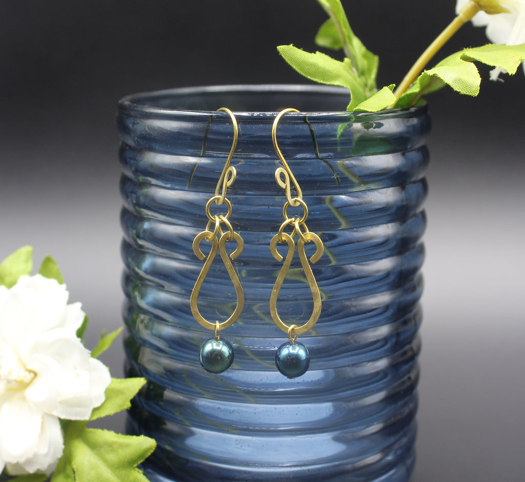 Hammered brass teardrop earrings with metallic dark teal bead drops in front of a blue glass vase and white flowers.