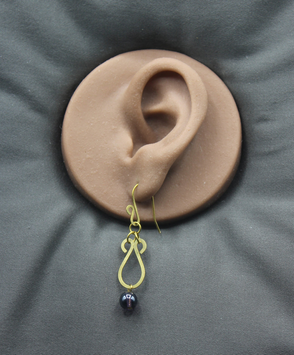 Hammered brass teardrop earrings with dark purple bead drops displayed on a clay model of an ear against a gray background