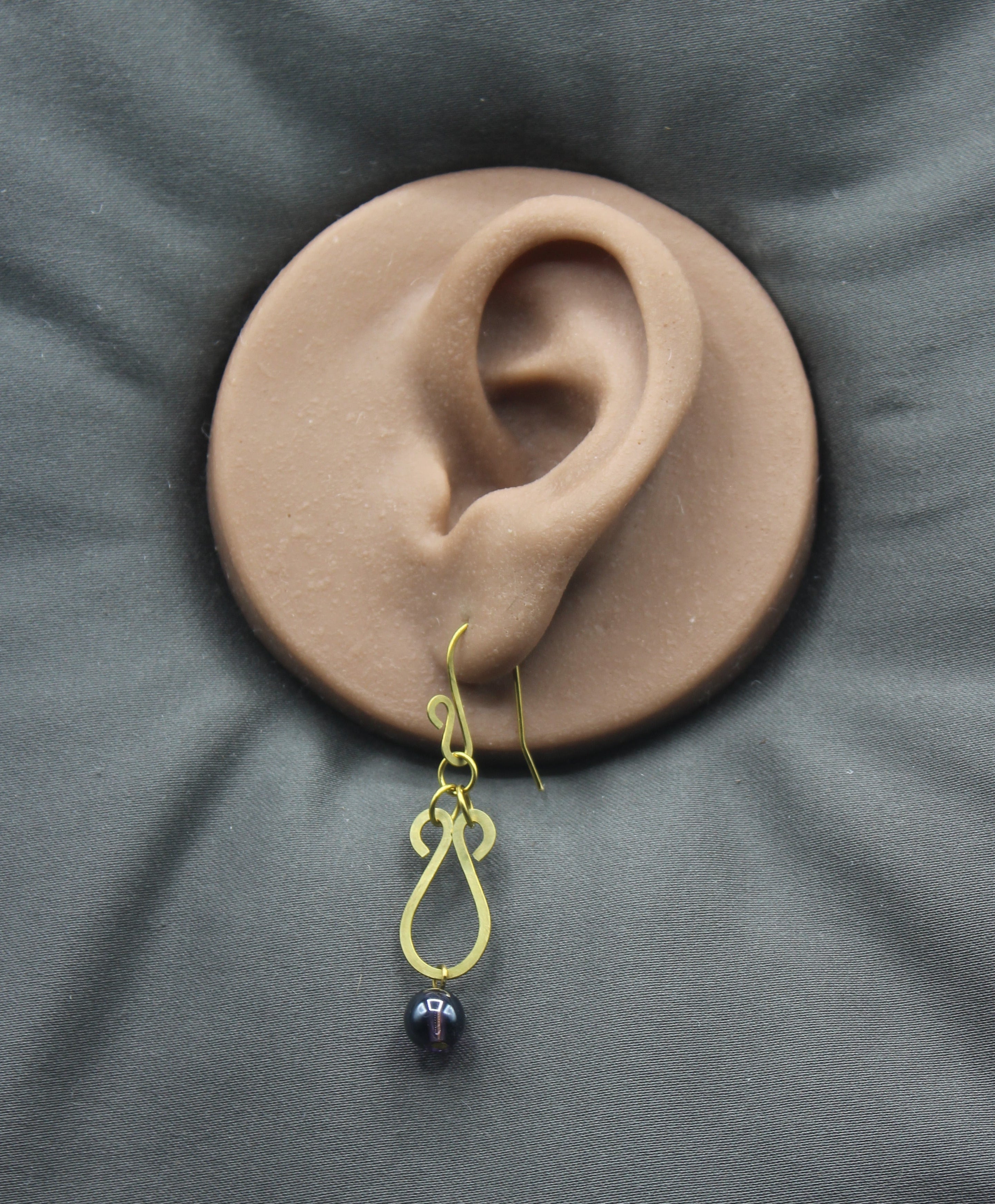 Hammered brass teardrop earrings with dark purple bead drops displayed on a clay model of an ear against a gray background