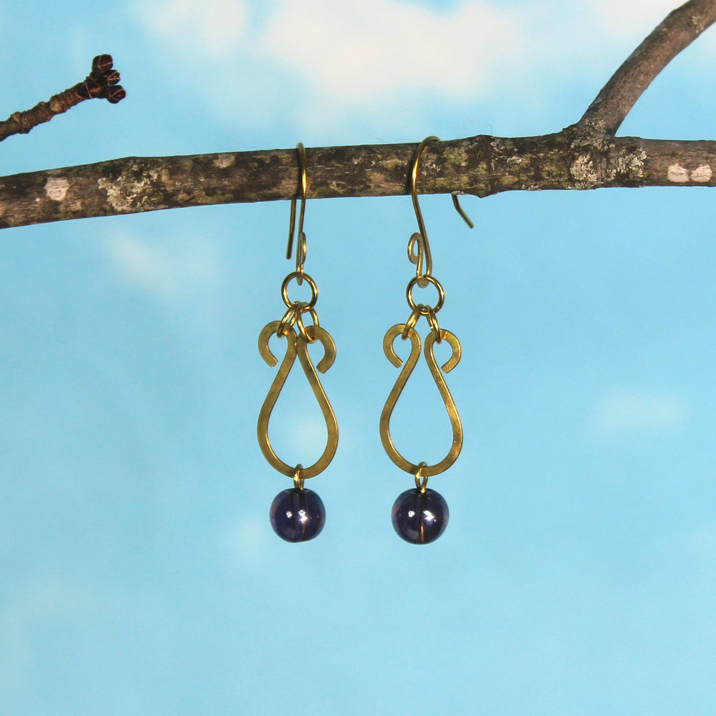 Hammered brass teardrop earrings with dark purple bead drops hanging from a branch against a blue sky.