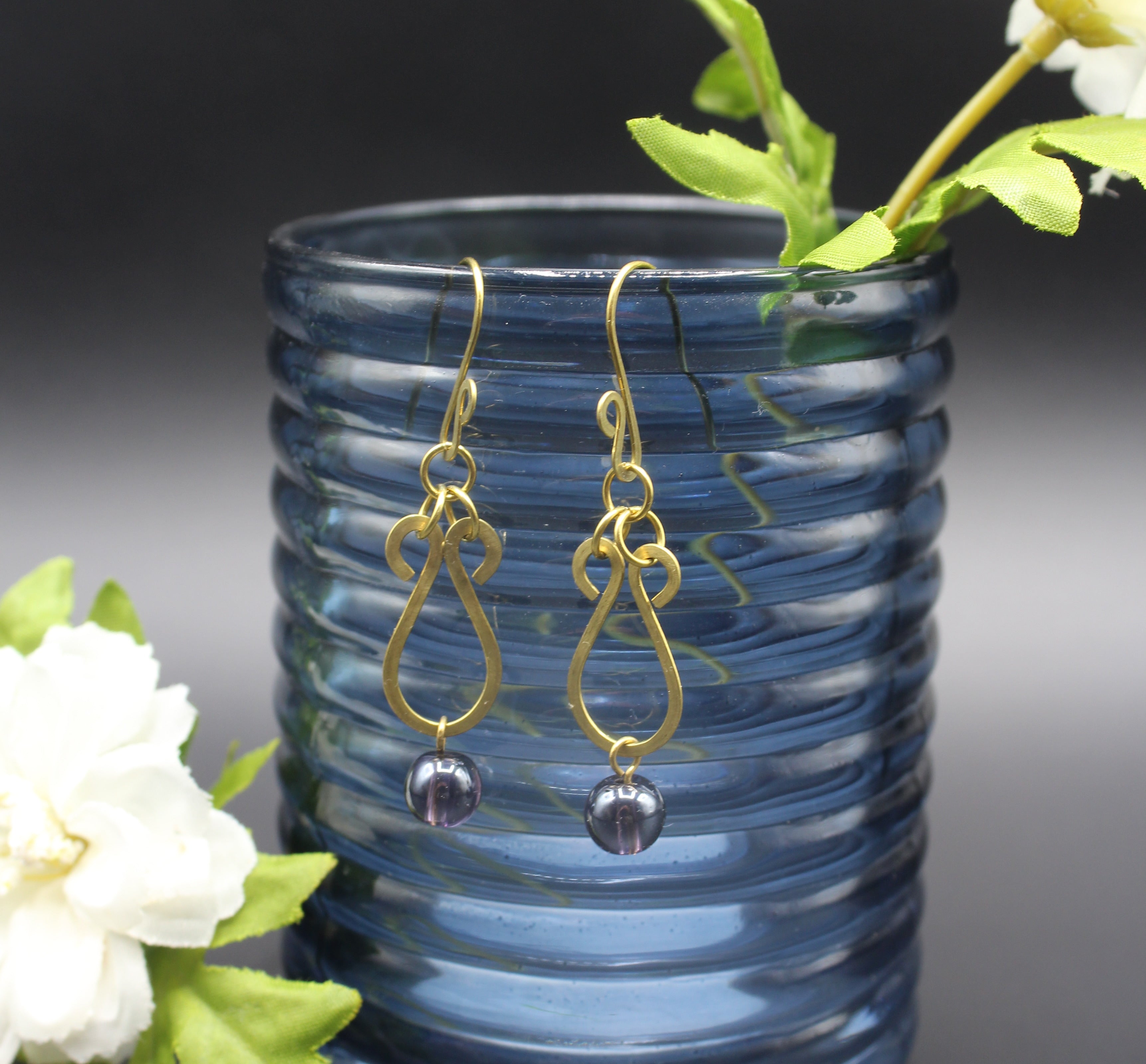 Hammered brass teardrop earrings with dark purple bead drops hanging from a blue glass vase.