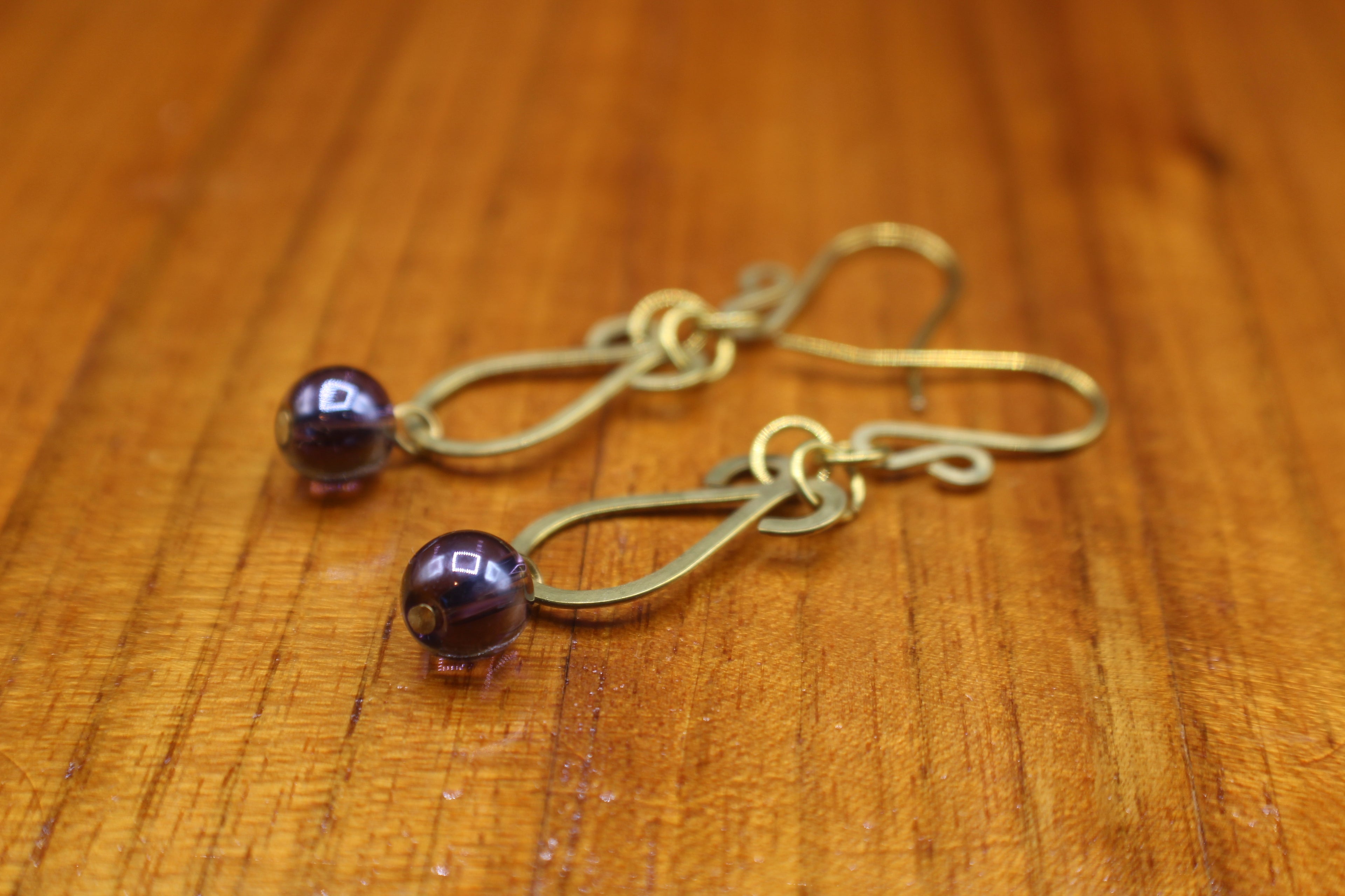 Hammered brass teardrop earrings with dark purple bead drops on a wooden surface