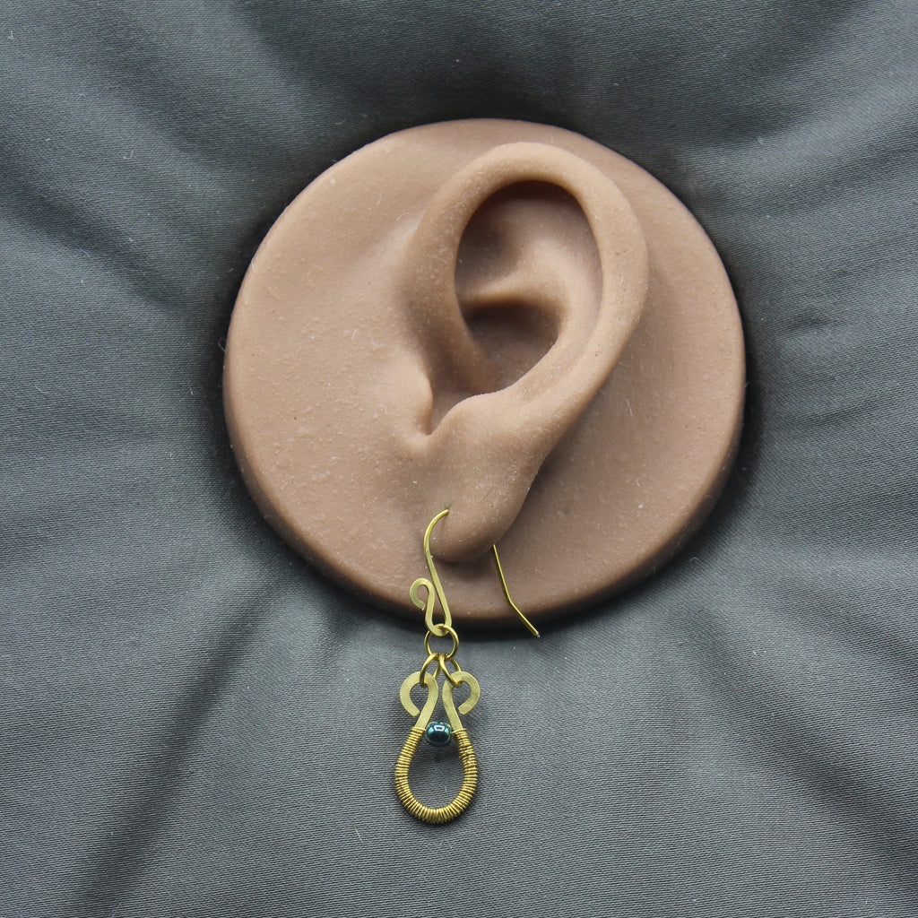 Hammered brass teardrop earrings wire-wrapped with metallic teal bead accents displayed on a model of a human ear.