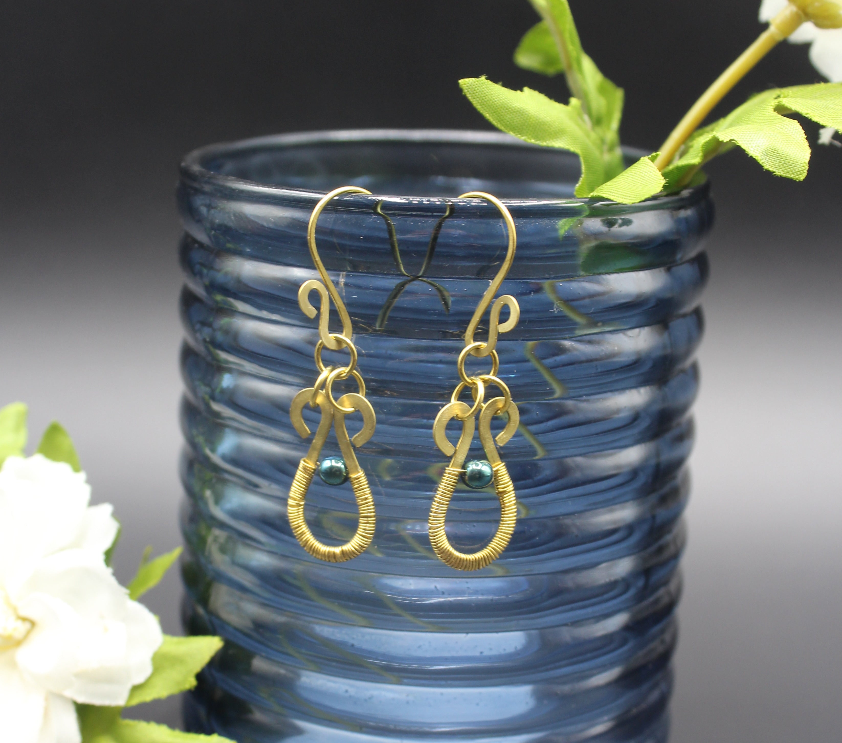 Hammered brass teardrop earrings wire-wrapped with metallic teal bead accents hanging from a blue glass vase with greenery.