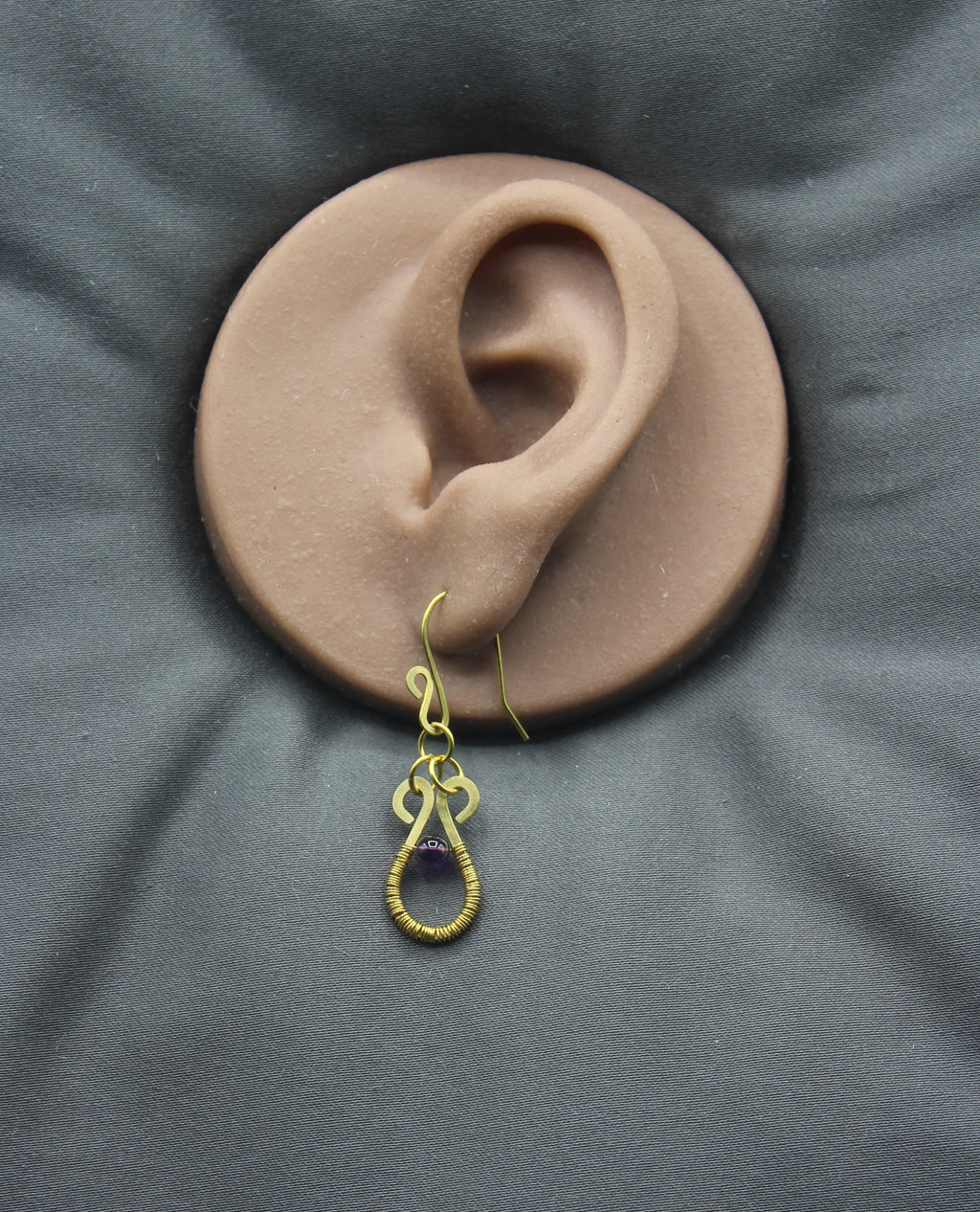 Hammered brass wire teardrop earring with purple amethyst gemstone and wire wrapping on a clay ear model against a gray background