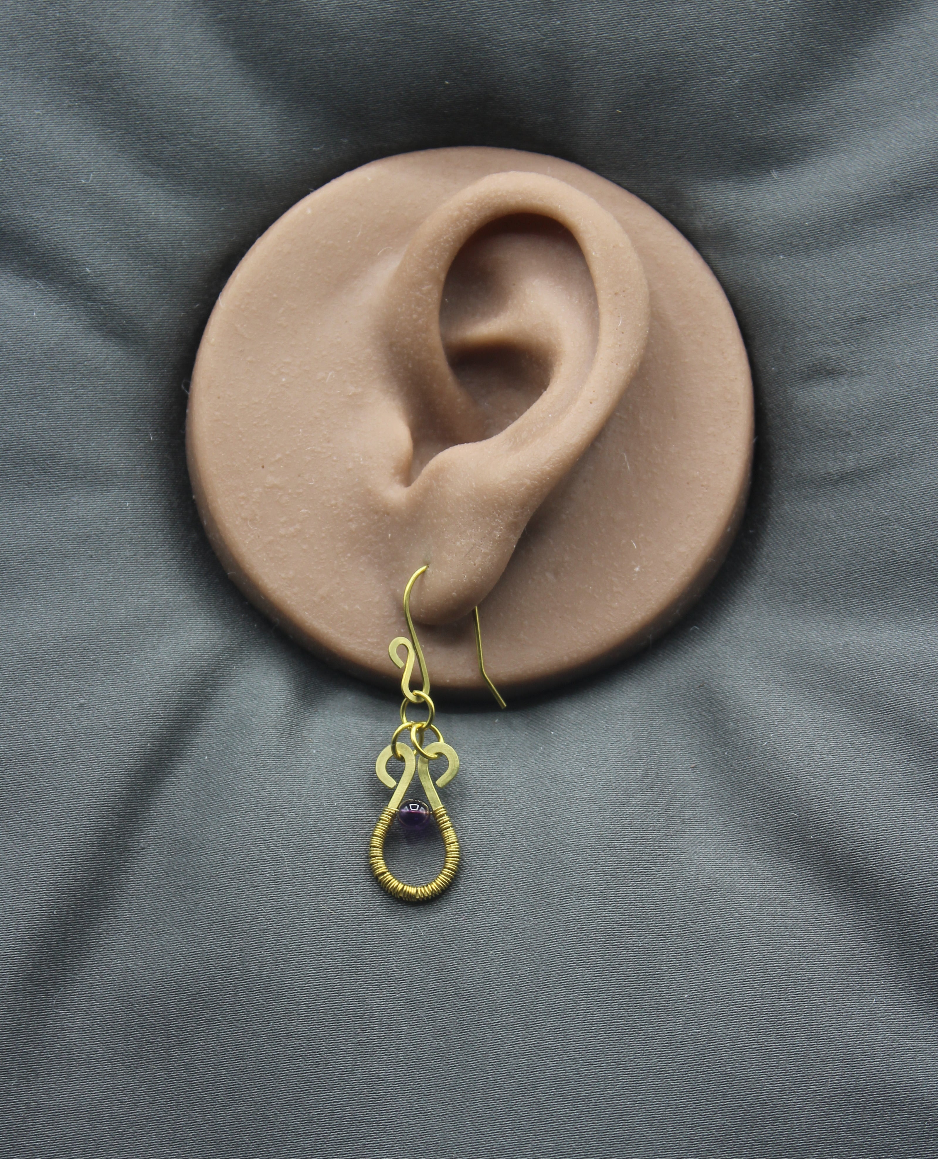 Hammered brass wire teardrop earring with purple amethyst gemstone and wire wrapping on a clay ear model against a gray background