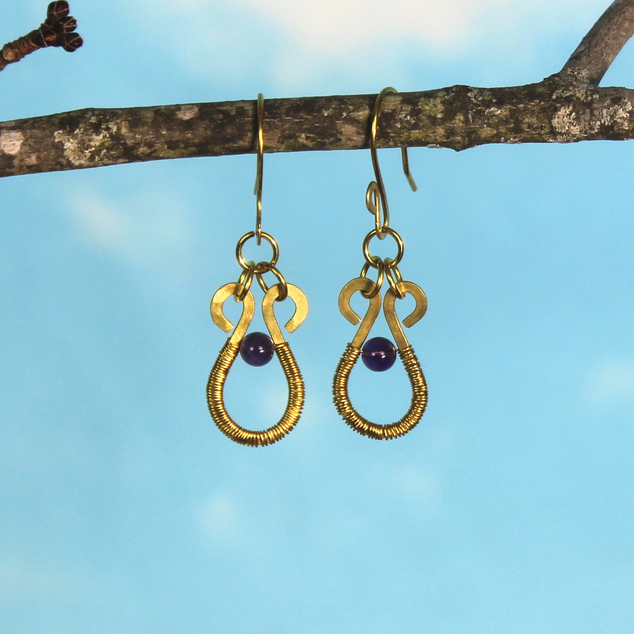 Brass hammered wire earrings with purple amethyst stones and wire wrapping hanging from a branch against a blue sky.