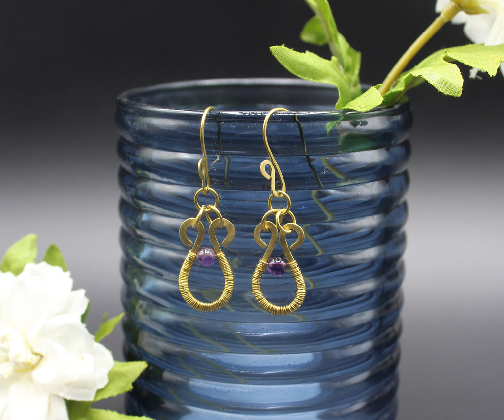 Hammered brass wire earrings with purple amethyst beads and wire wrapping, hanging from a blue glass vase with flowers.