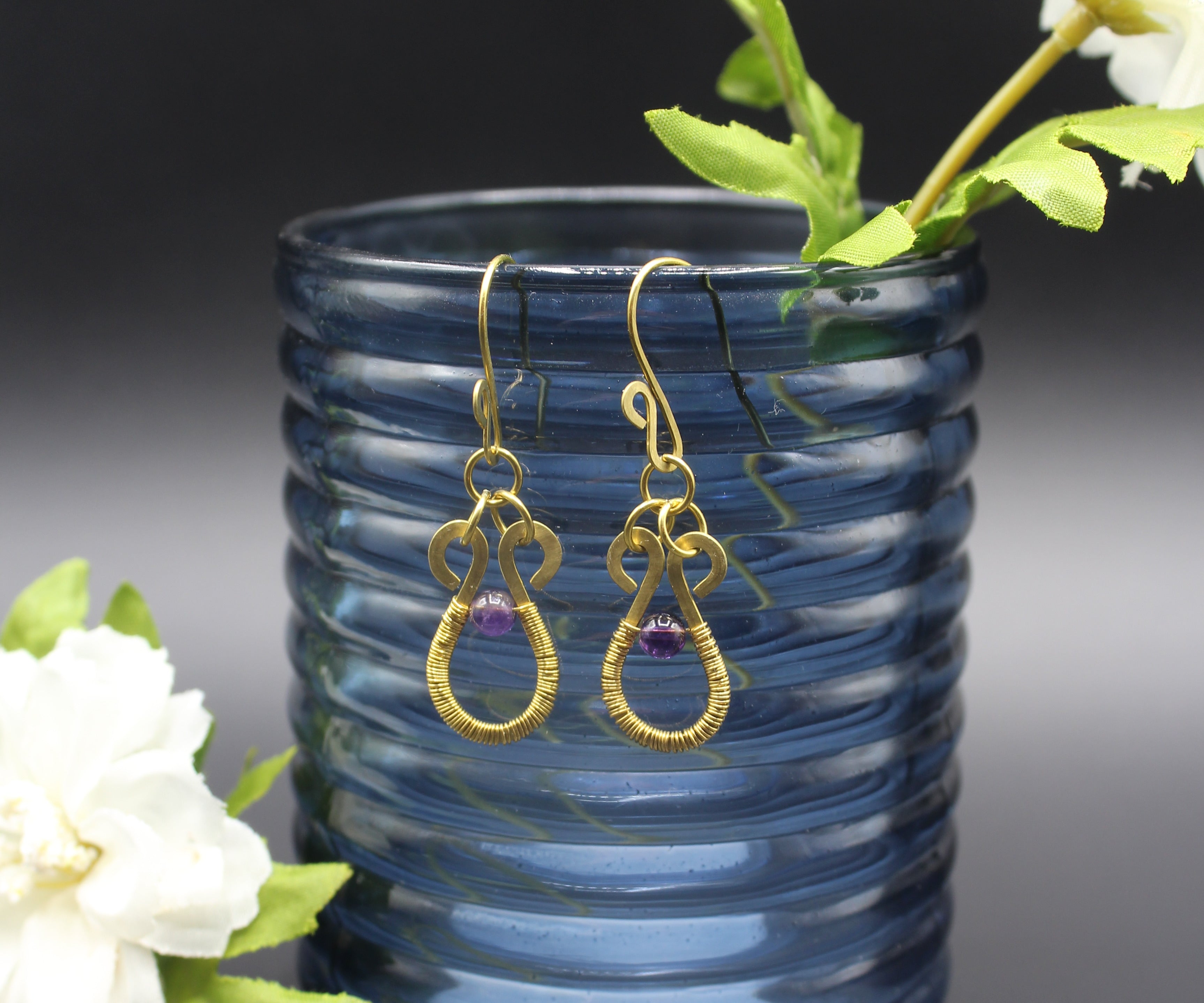 Hammered brass wire earrings with purple amethyst beads and wire wrapping, hanging from a blue glass vase with flowers.