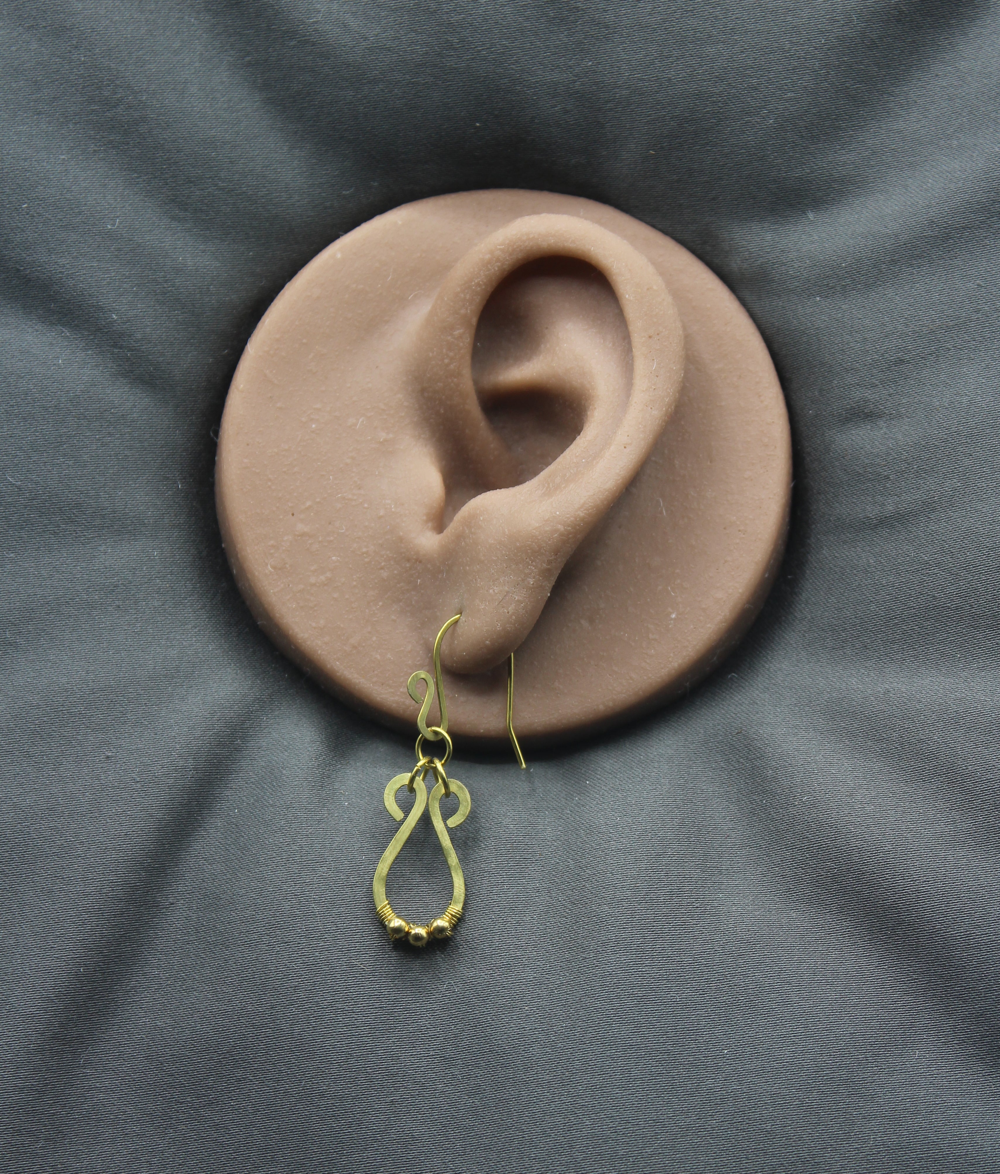 Hammered brass teardrop earrings with small brass bead accents on a clay model of an ear on a gray fabric background