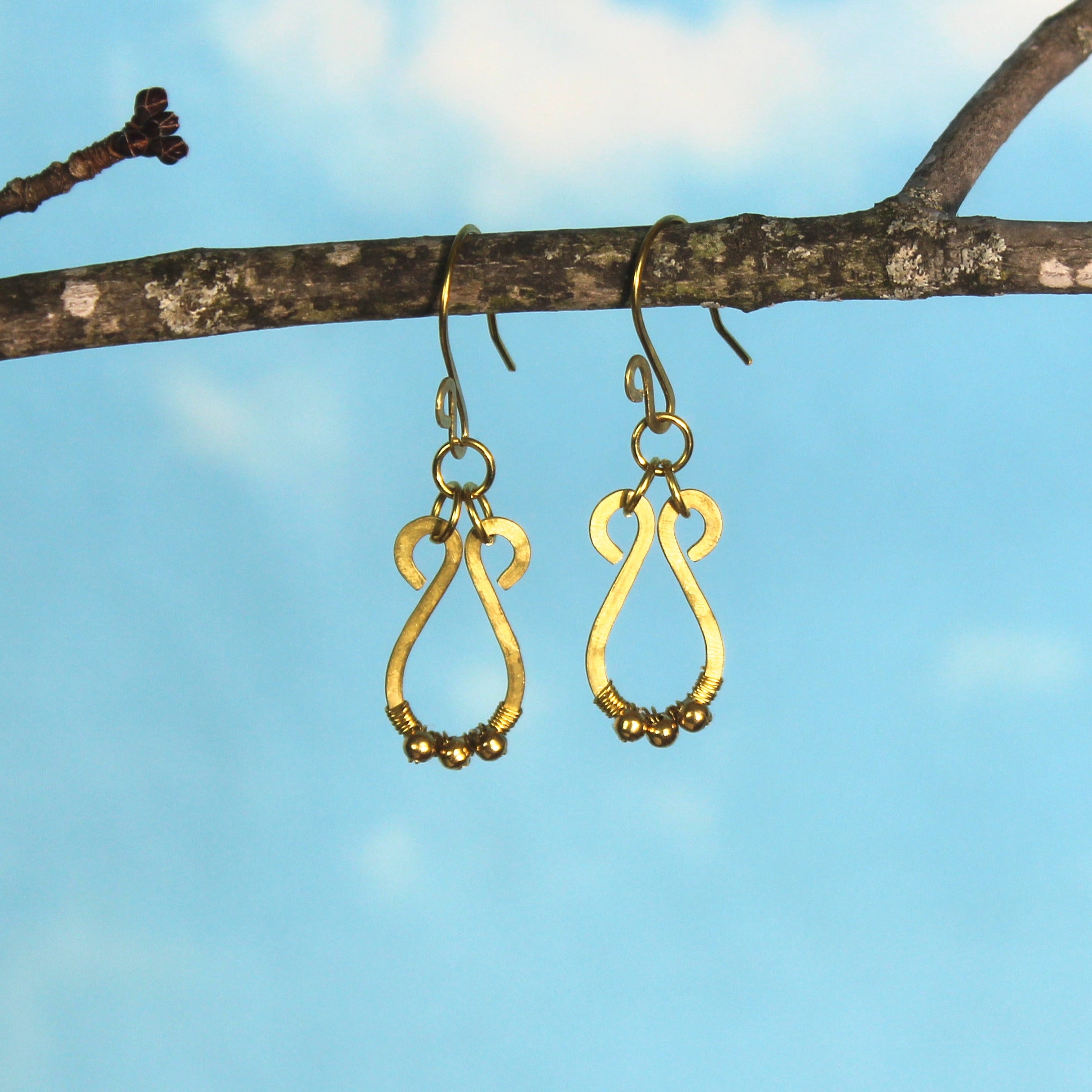 Hammered brass teardrop earrings with small brass bead accents hanging from a branch against a blue sky background