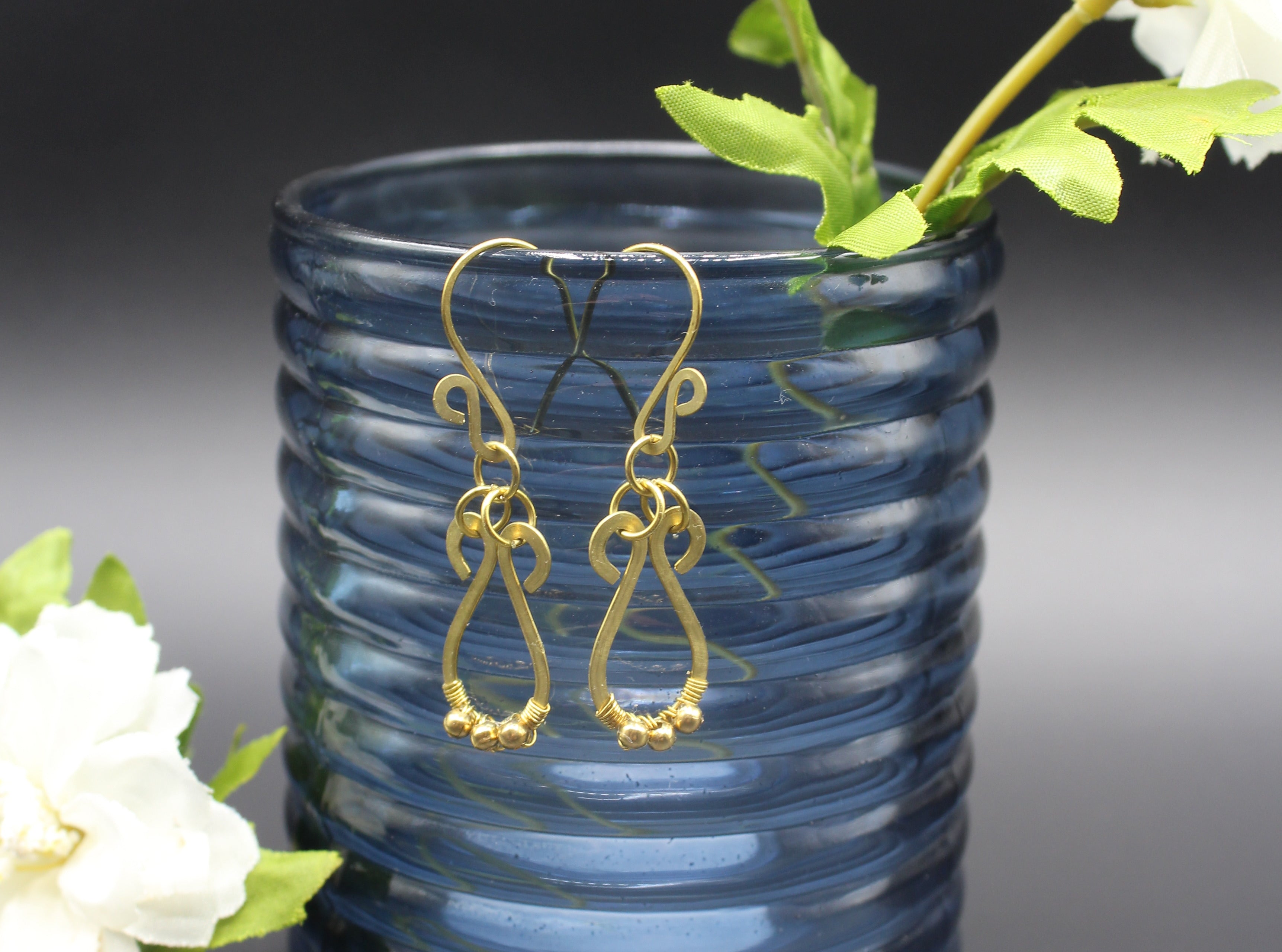 Hammered brass teardrop earrings with small brass bead accents hanging from a blue glass vase with greenery and flowers.