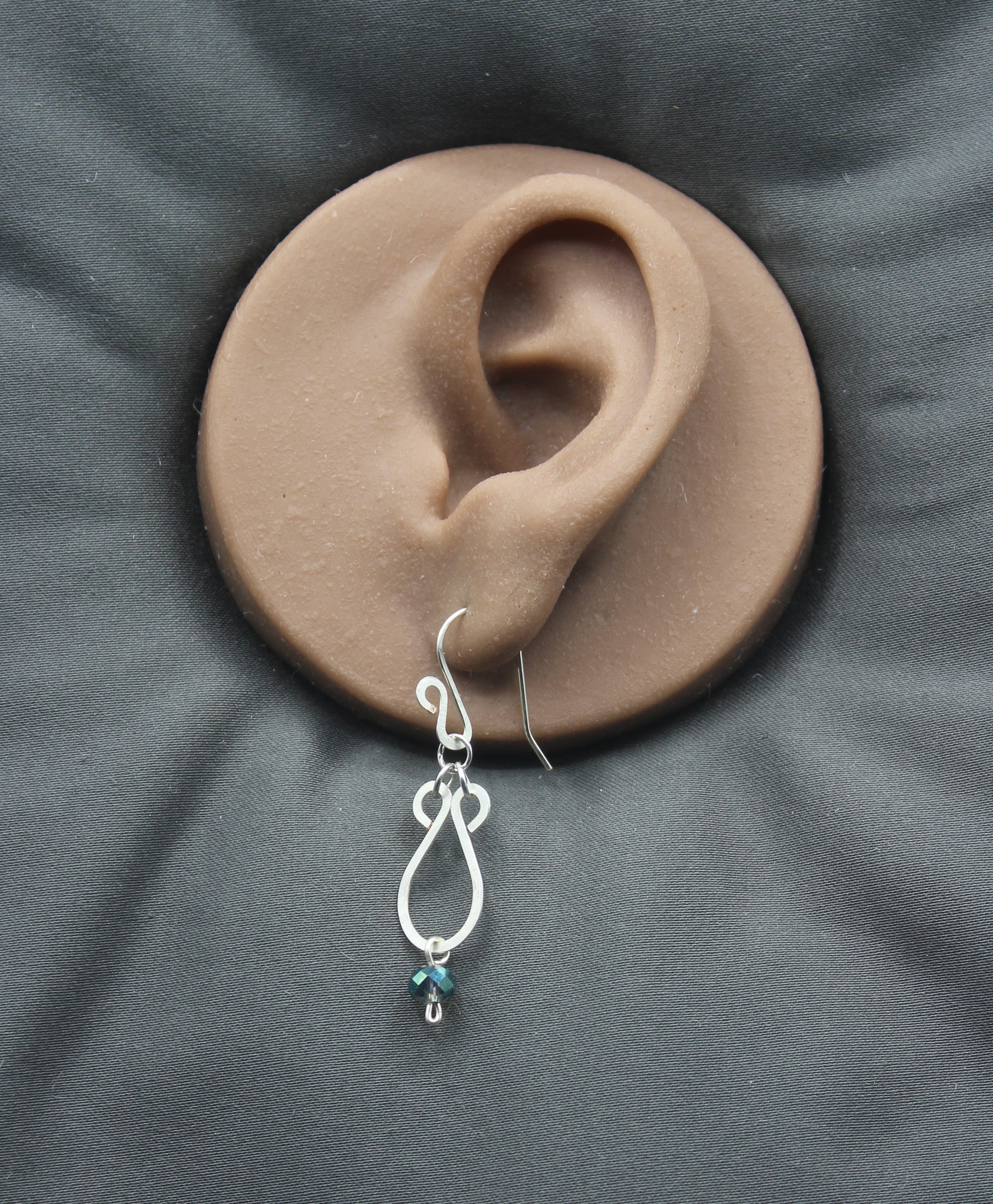 Hammered silver teardrop earrings with faceted blue bead drops on a clay model of an ear against a gray background