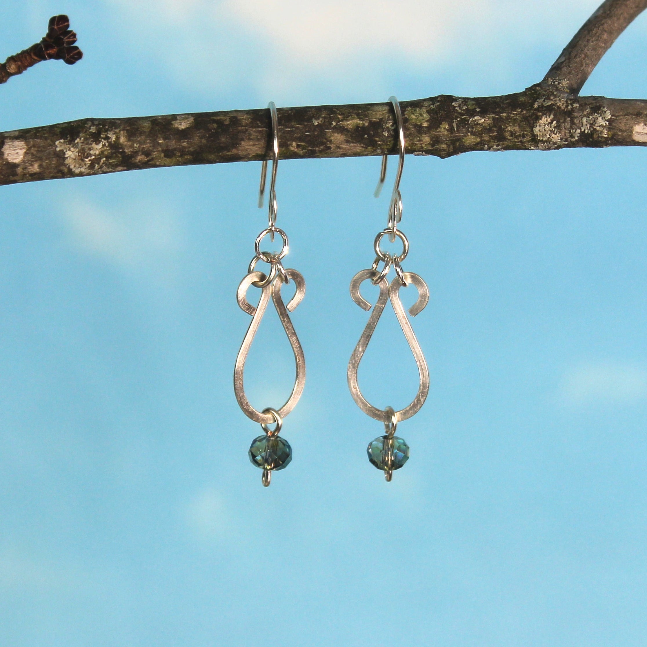 Hammered silver teardrop earrings with faceted blue bead drops hanging on a branch against a blue sky.