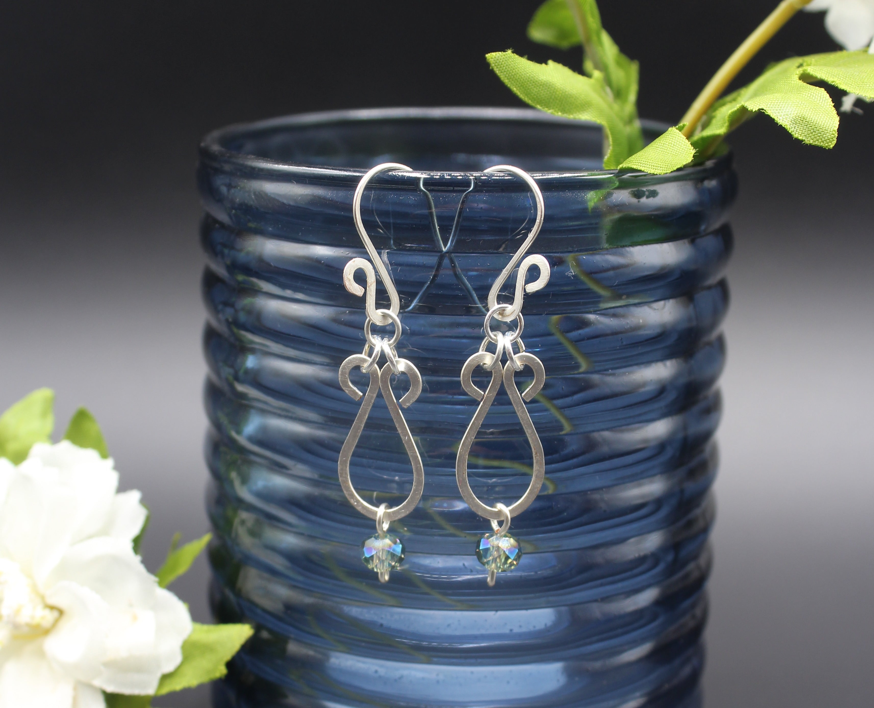 Hammered silver teardrop earrings with faceted blue bead drops hanging from a blue glass vase.