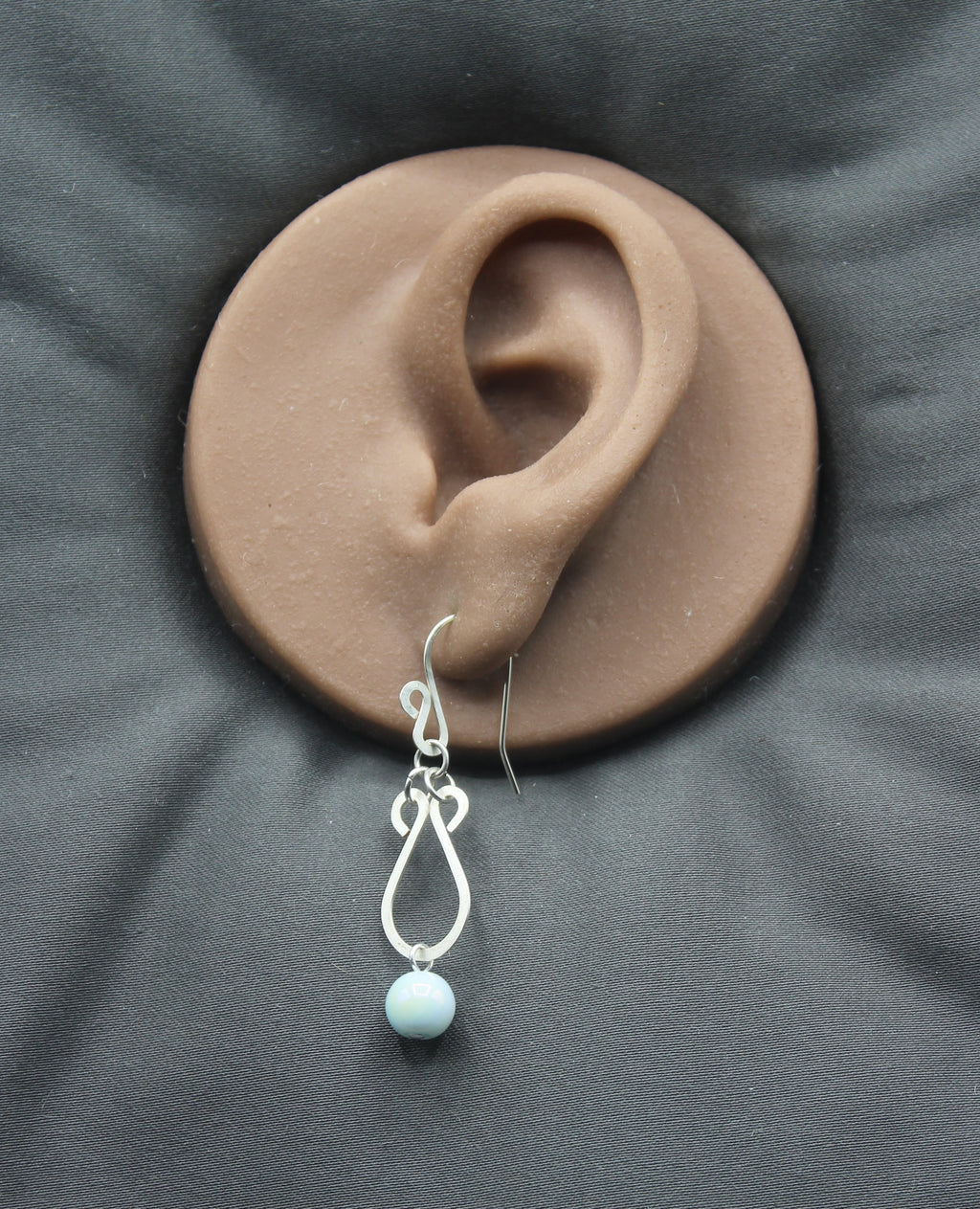 Hammered silver earring with a pale blue bead drop on a clay model of an ear