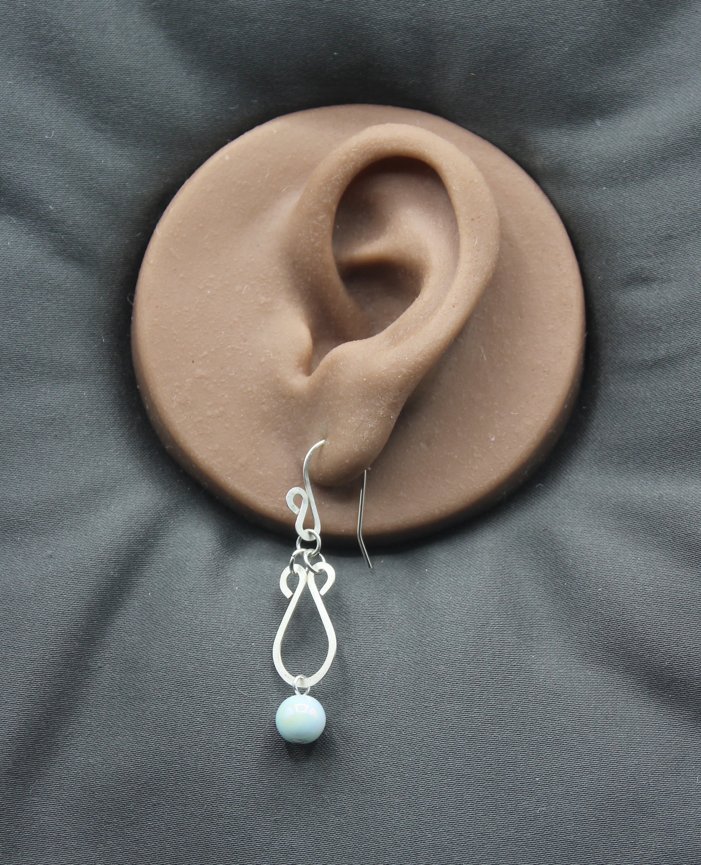 Hammered silver earring with a pale blue bead drop on a clay model of an ear