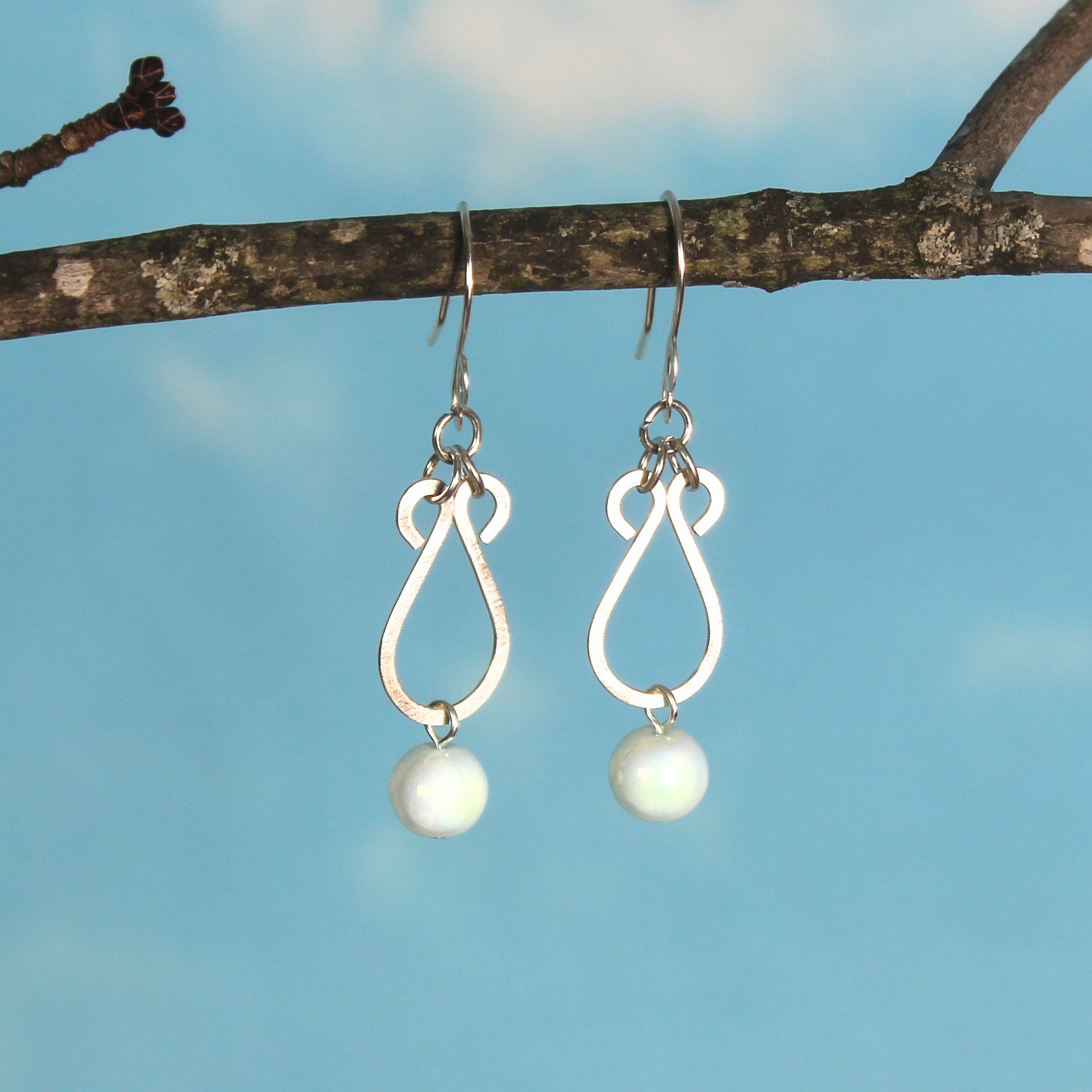Hammered silver earrings with pale blue bead drops hanging from a branch against a blue sky background