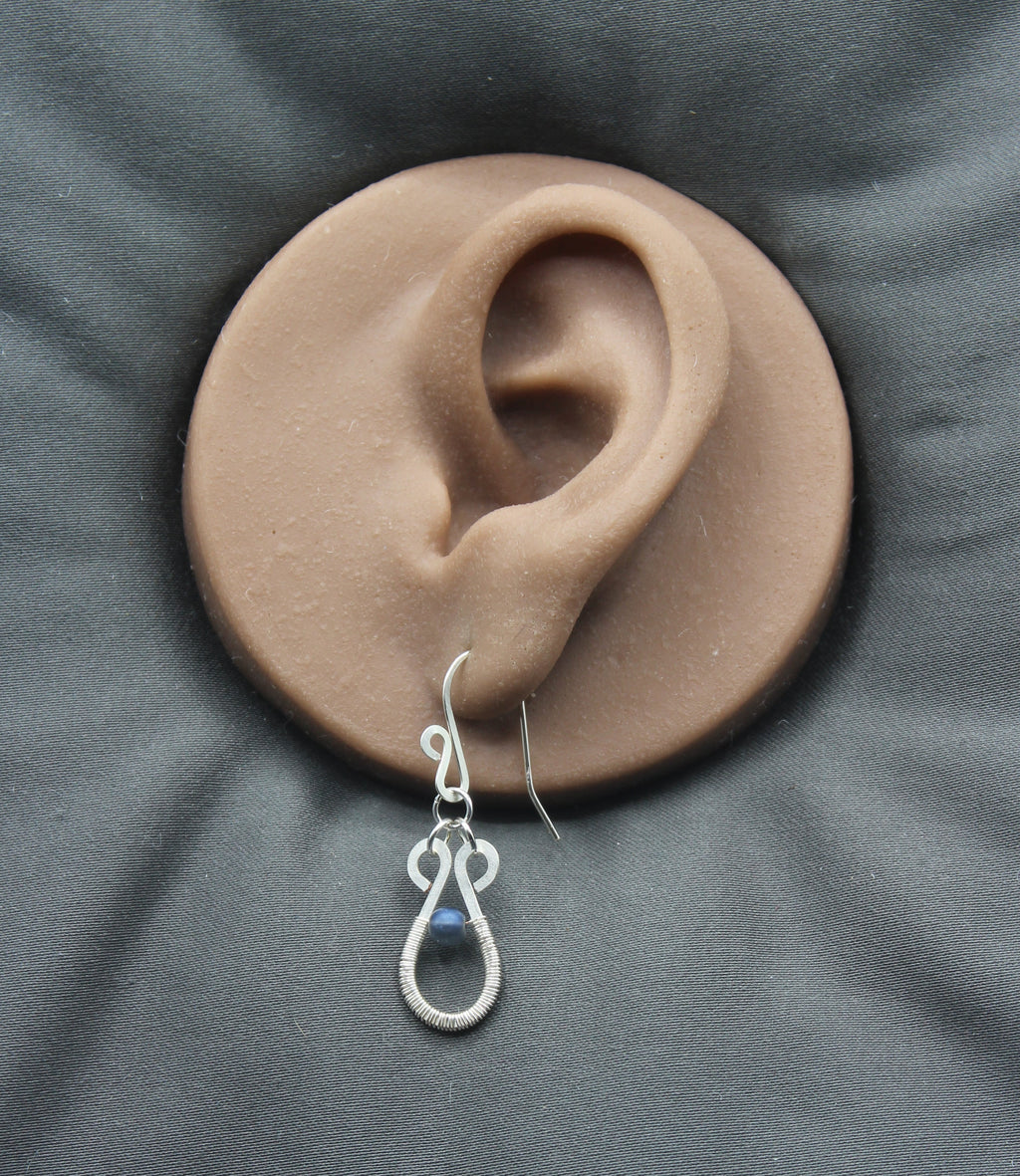 Hammered silver teardrop earring with a wire-wrapped blue bead accent on a clay model of an ear with a gray background