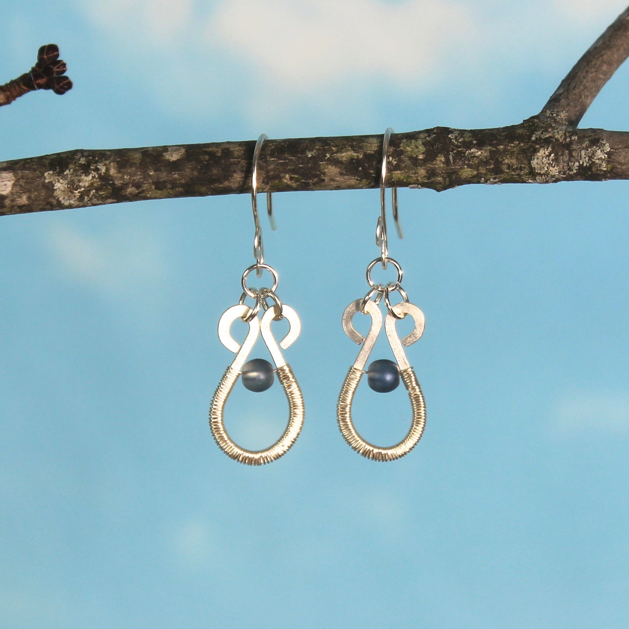 Hammered silver teardrop earrings with wire-wrapped blue bead accents hanging on a branch against a blue sky.
