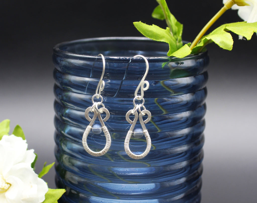 Hammered silver teardrop earrings with wire-wrapped blue bead accents hanging from a blue vase.