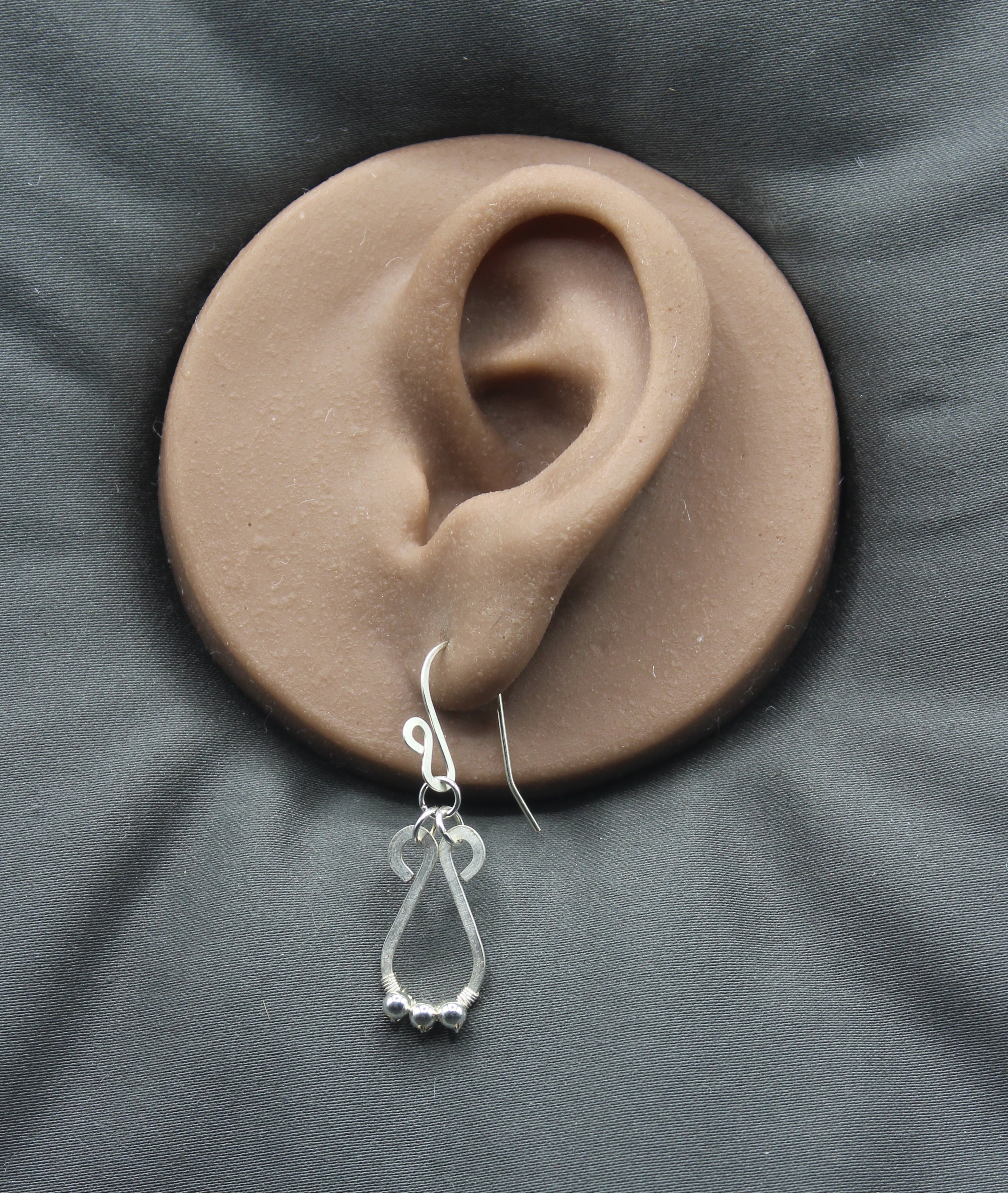 Hammered silver teardrop earring with small silver bead accents hanging from a clay ear model on a gray background