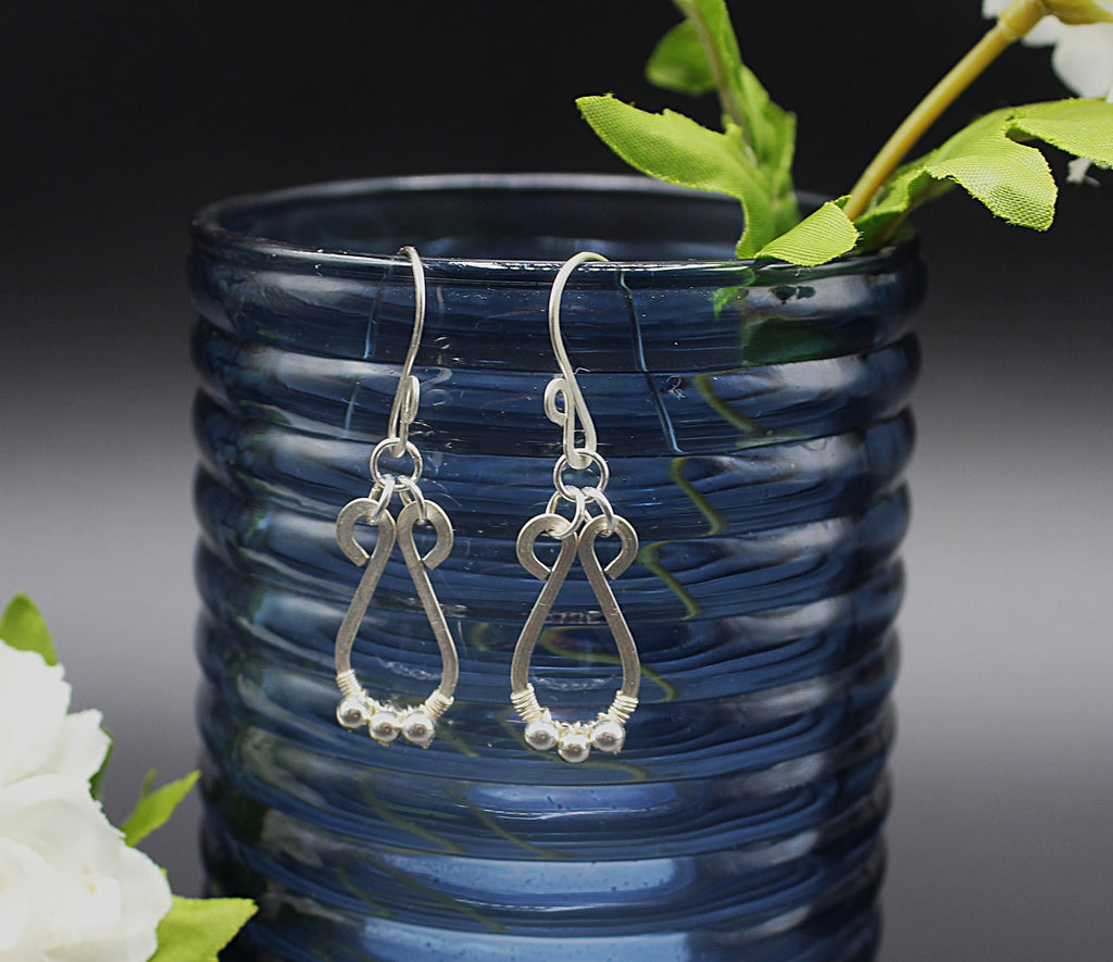 Hammered silver teardrop earrings with small silver bead accents hanging from a blue vase.