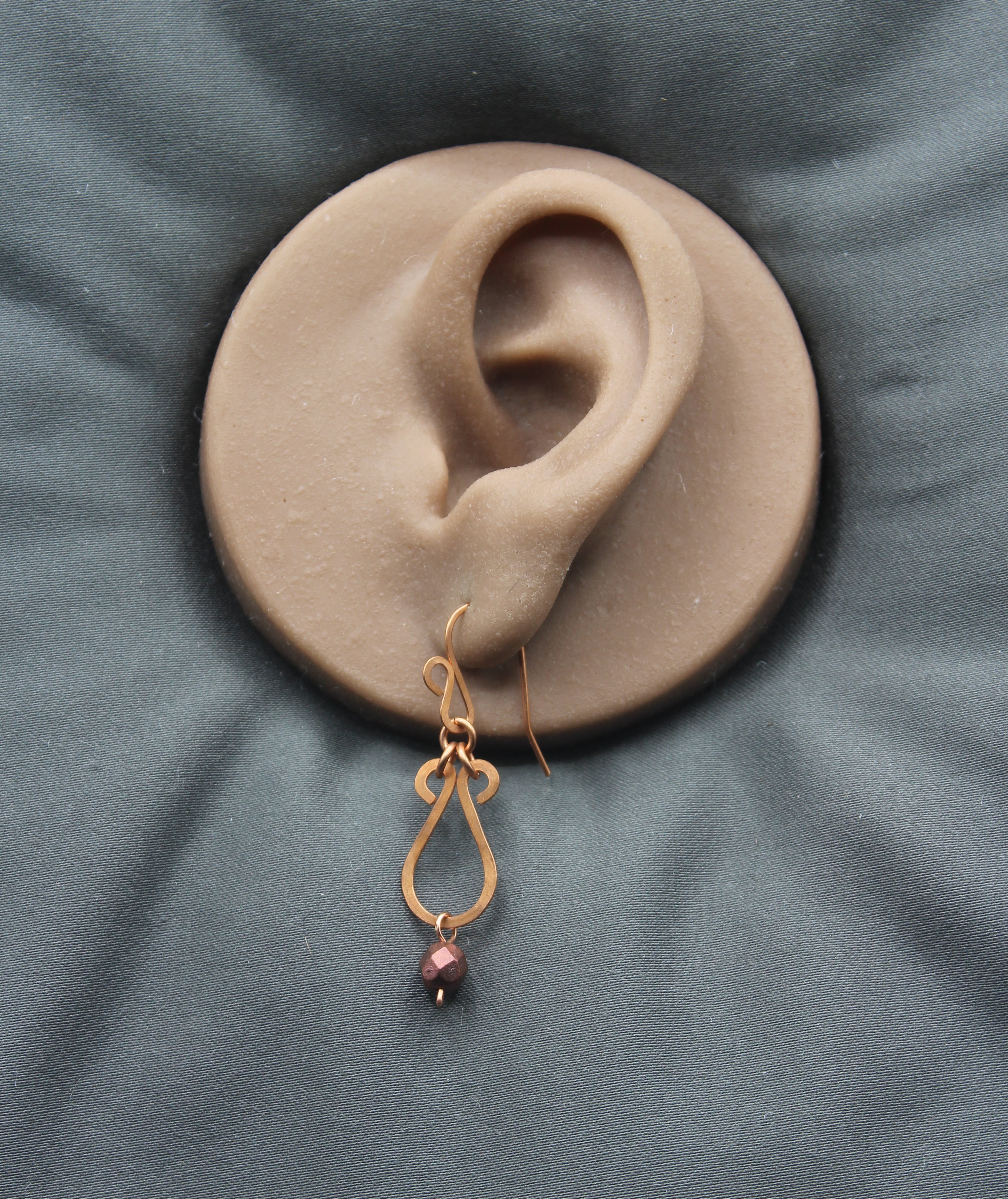 Hammered copper teardrop earrings with metallic finish, faceted glass beads on a clay model of an ear
