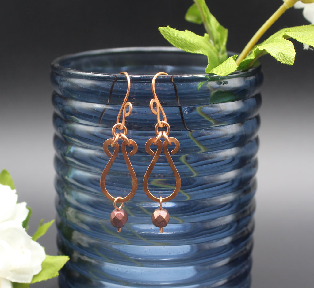 Hammered copper teardrop earrings with metallic finish, faceted glass beads hanging in front of a blue glass vase.