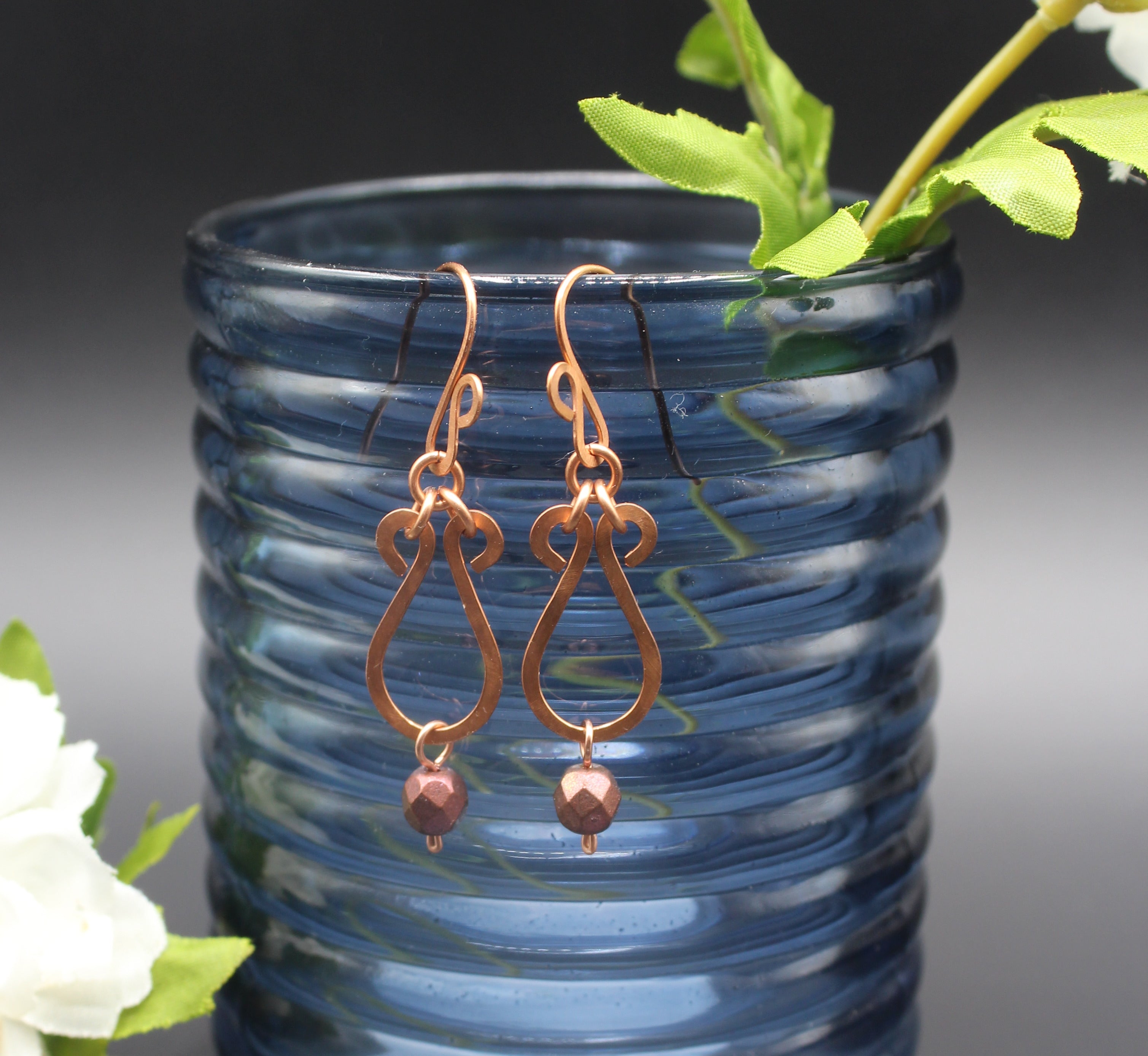 Hammered copper teardrop earrings with metallic finish, faceted glass beads hanging in front of a blue glass vase.