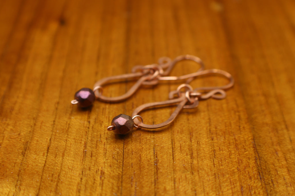Hammered copper teardrop earrings with metallic finish, faceted glass beads on a wooden surface