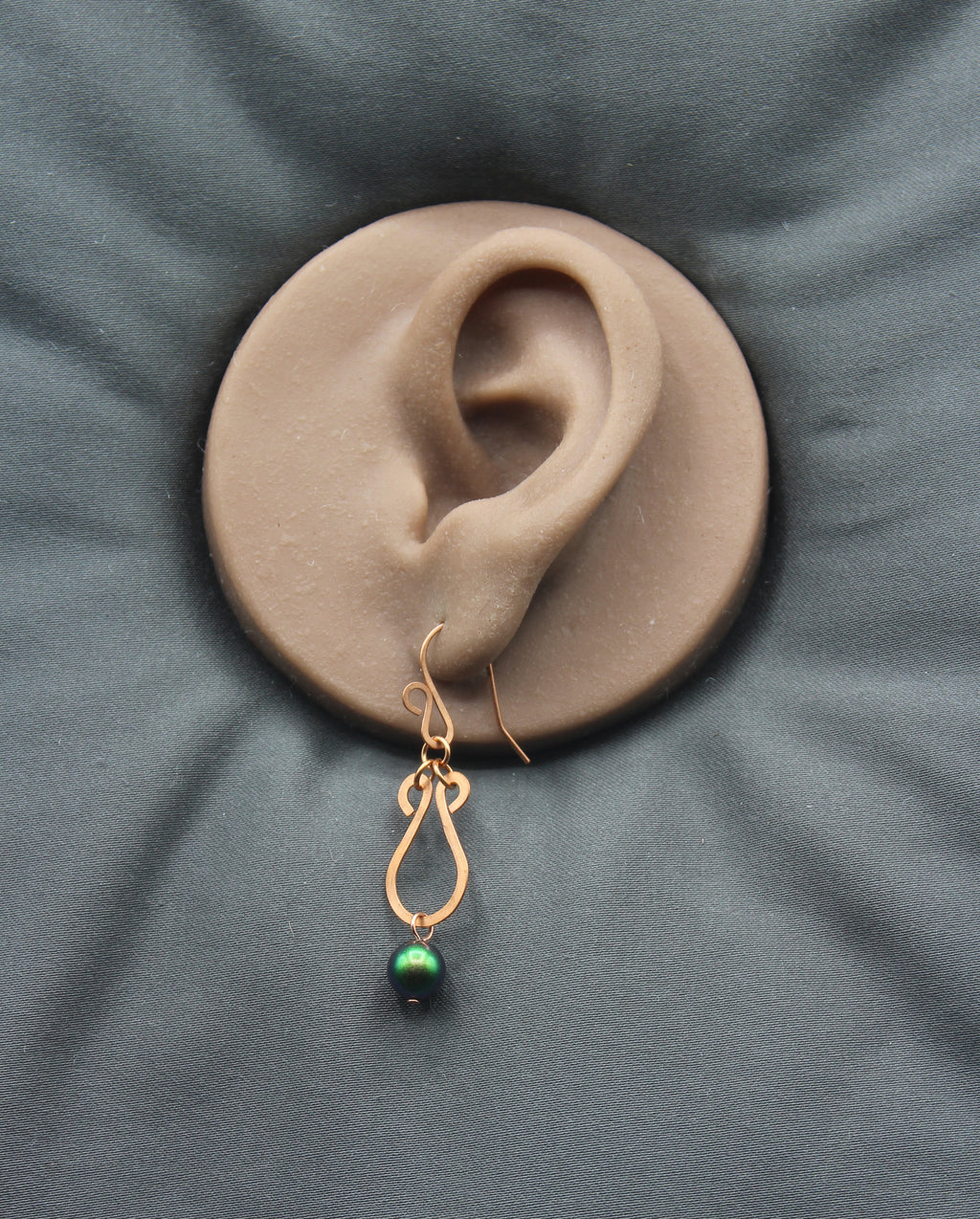 Copper teardrop earring with a green bead on a clay model of an ear