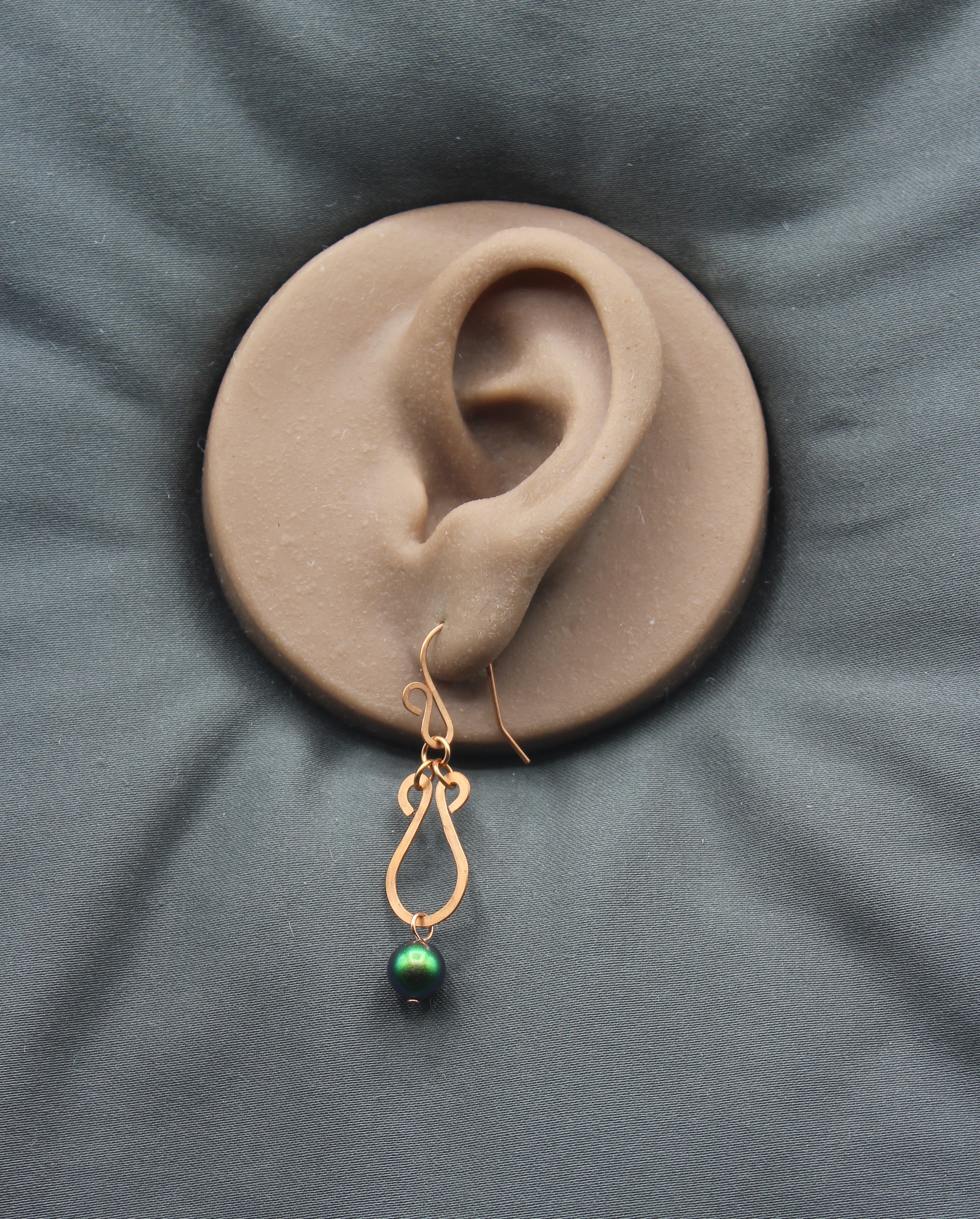 Copper teardrop earring with a green bead on a clay model of an ear