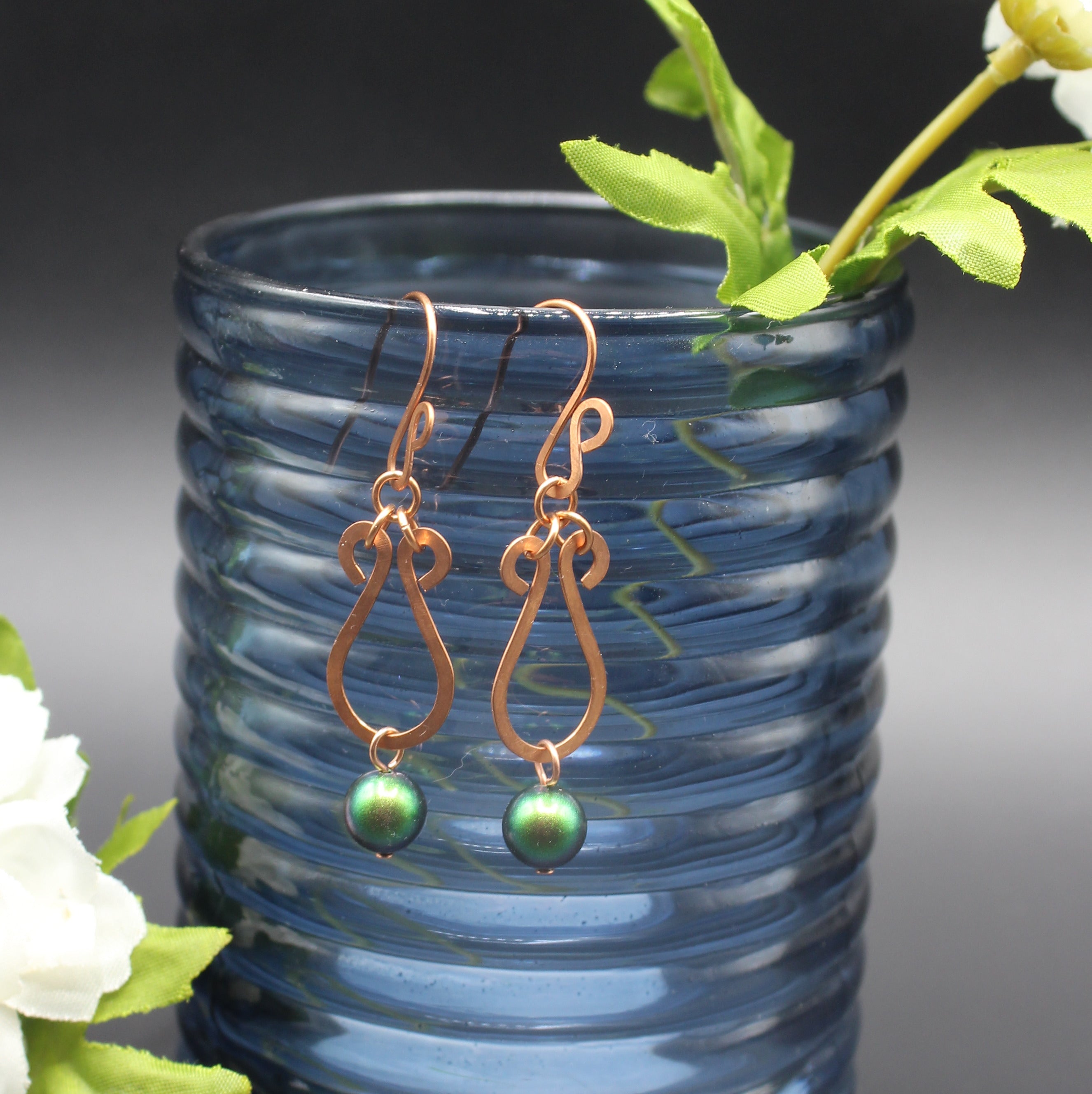 Hammered copper teardrop earrings with green beads in front of a blue glass vase.