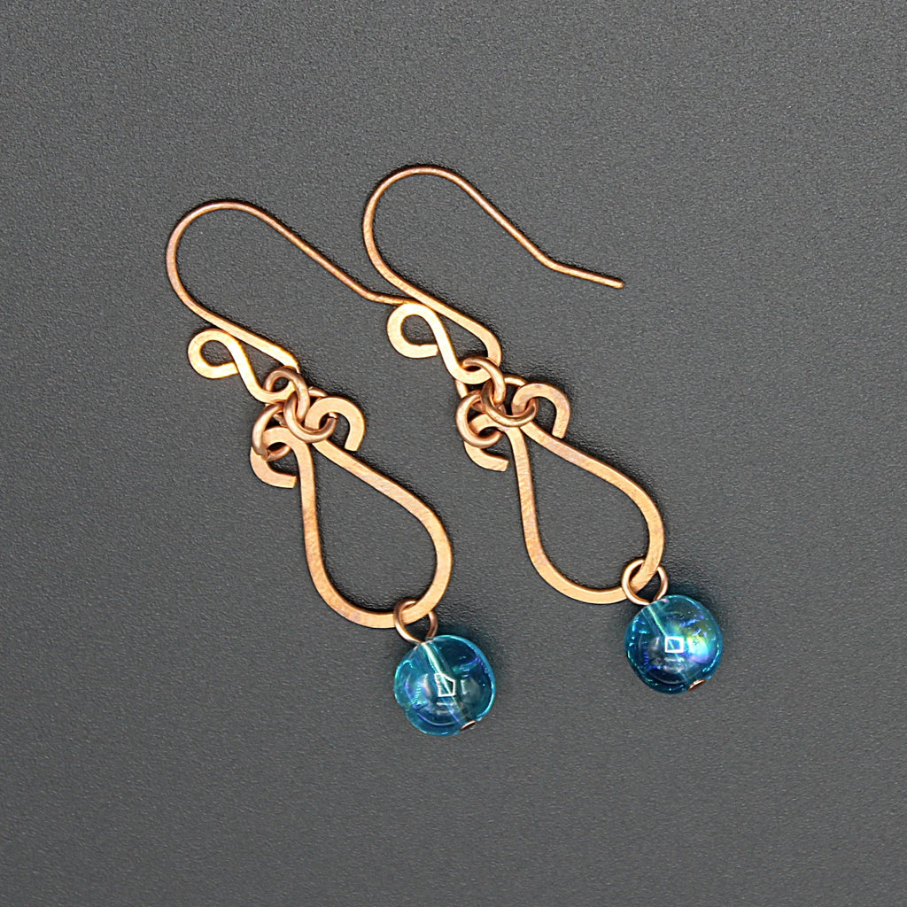 Hammered copper teardrop earrings with blue transparent bead drops on a gray background.