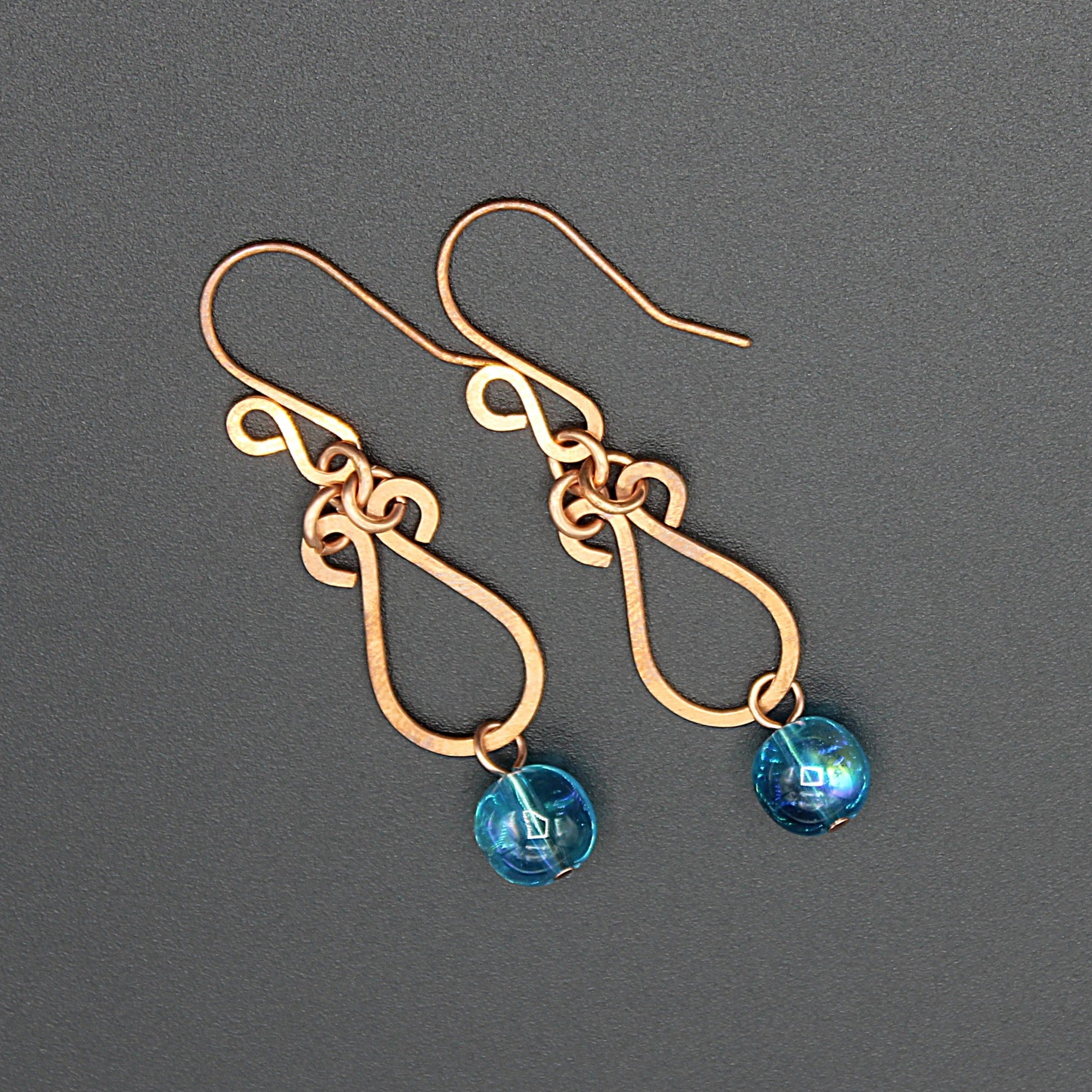Hammered copper teardrop earrings with blue transparent bead drops on a gray background.