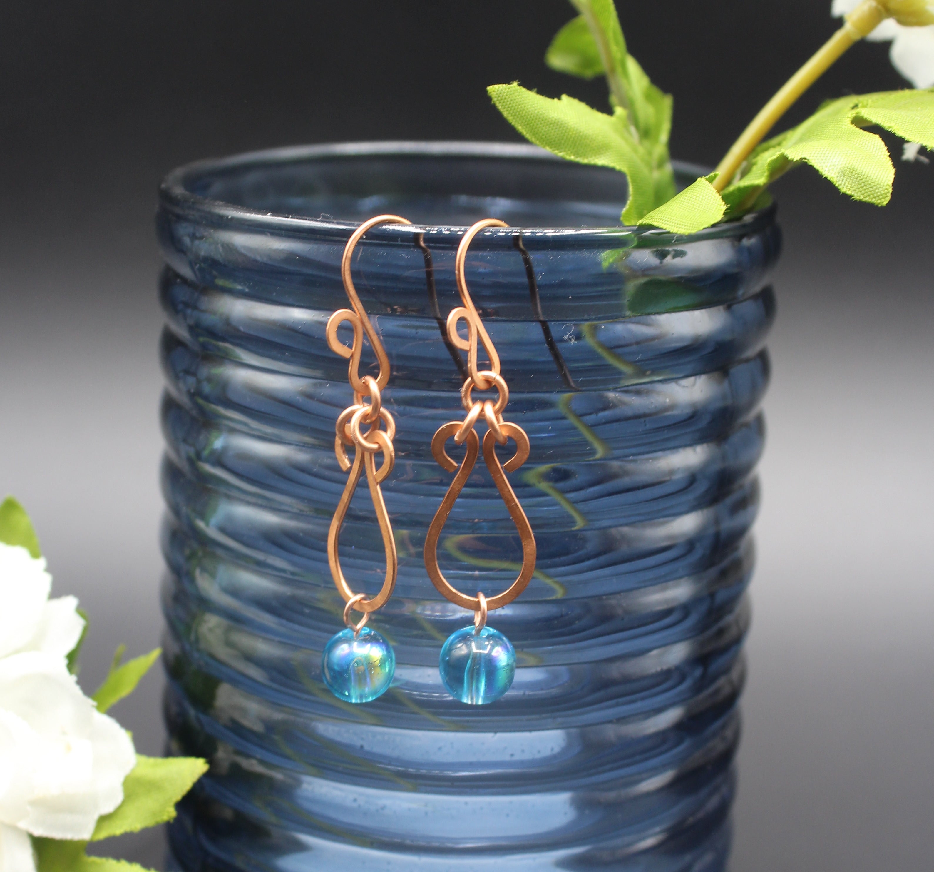 Hammered copper teardrop earrings with blue transparent bead drops in front of a blue glass vase.