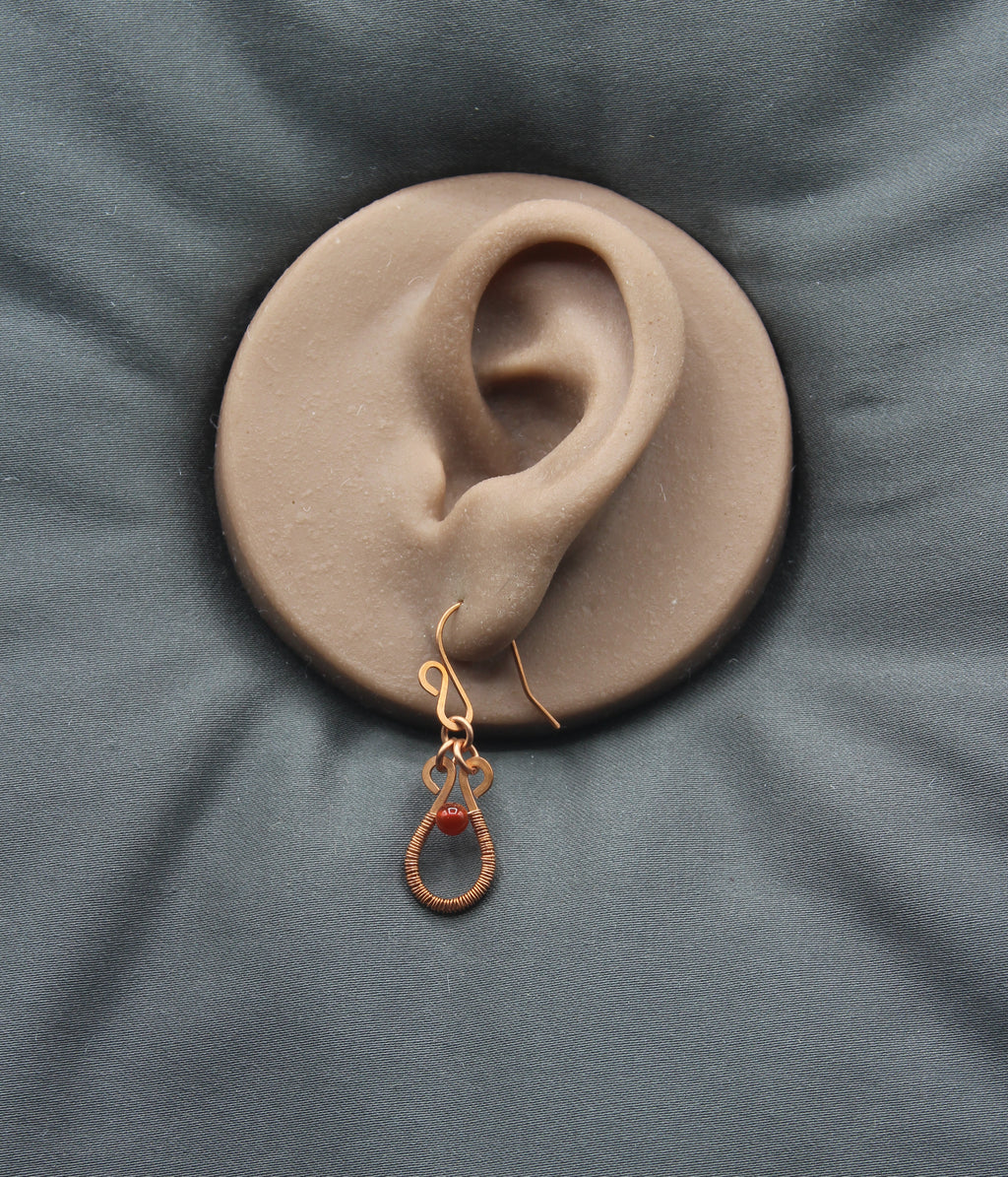 Flat wire copper teardrop earring wire wrapped with a red carnelian bead accent on a clay model of an ear against a gray background