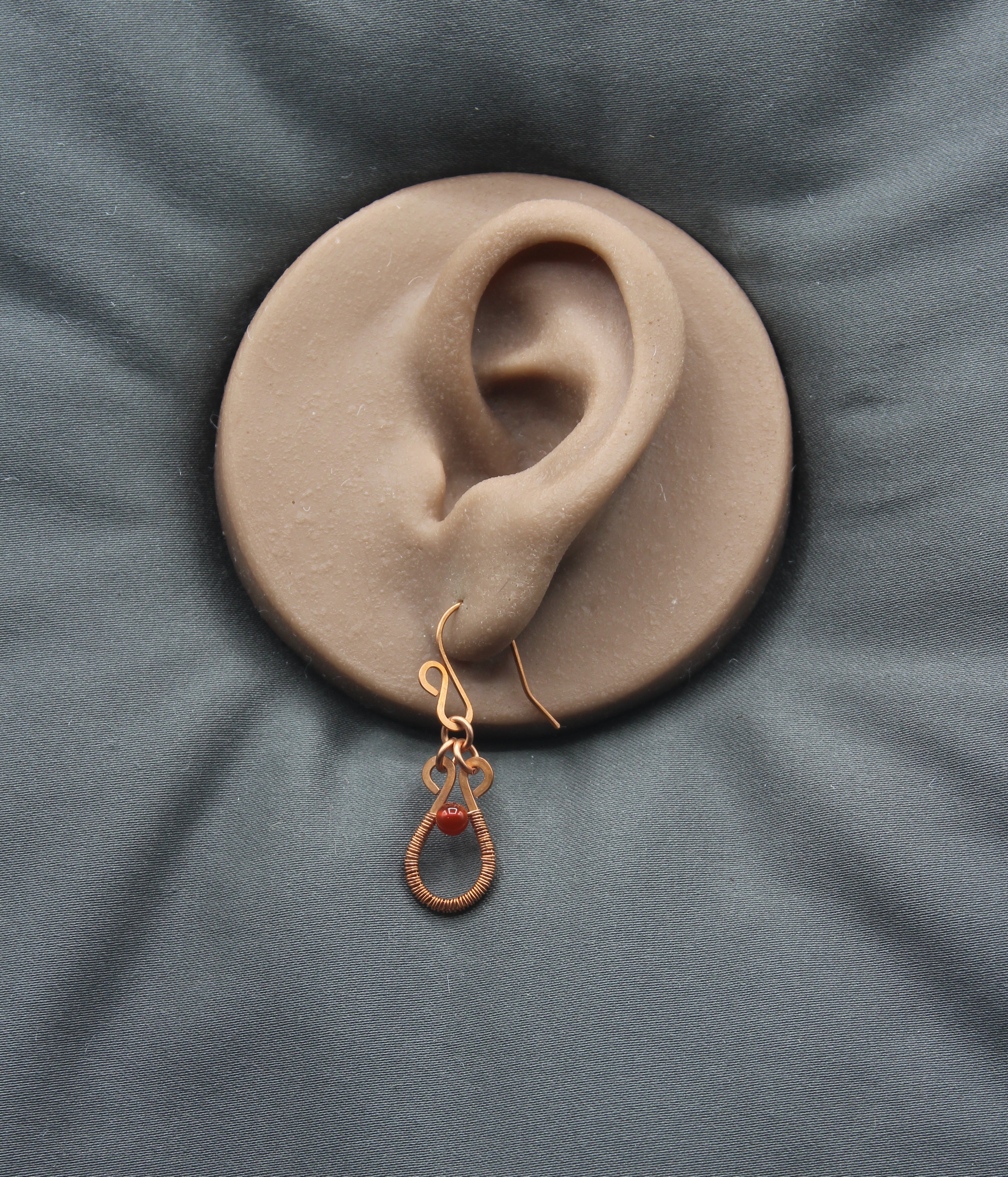Flat wire copper teardrop earring wire wrapped with a red carnelian bead accent on a clay model of an ear against a gray background