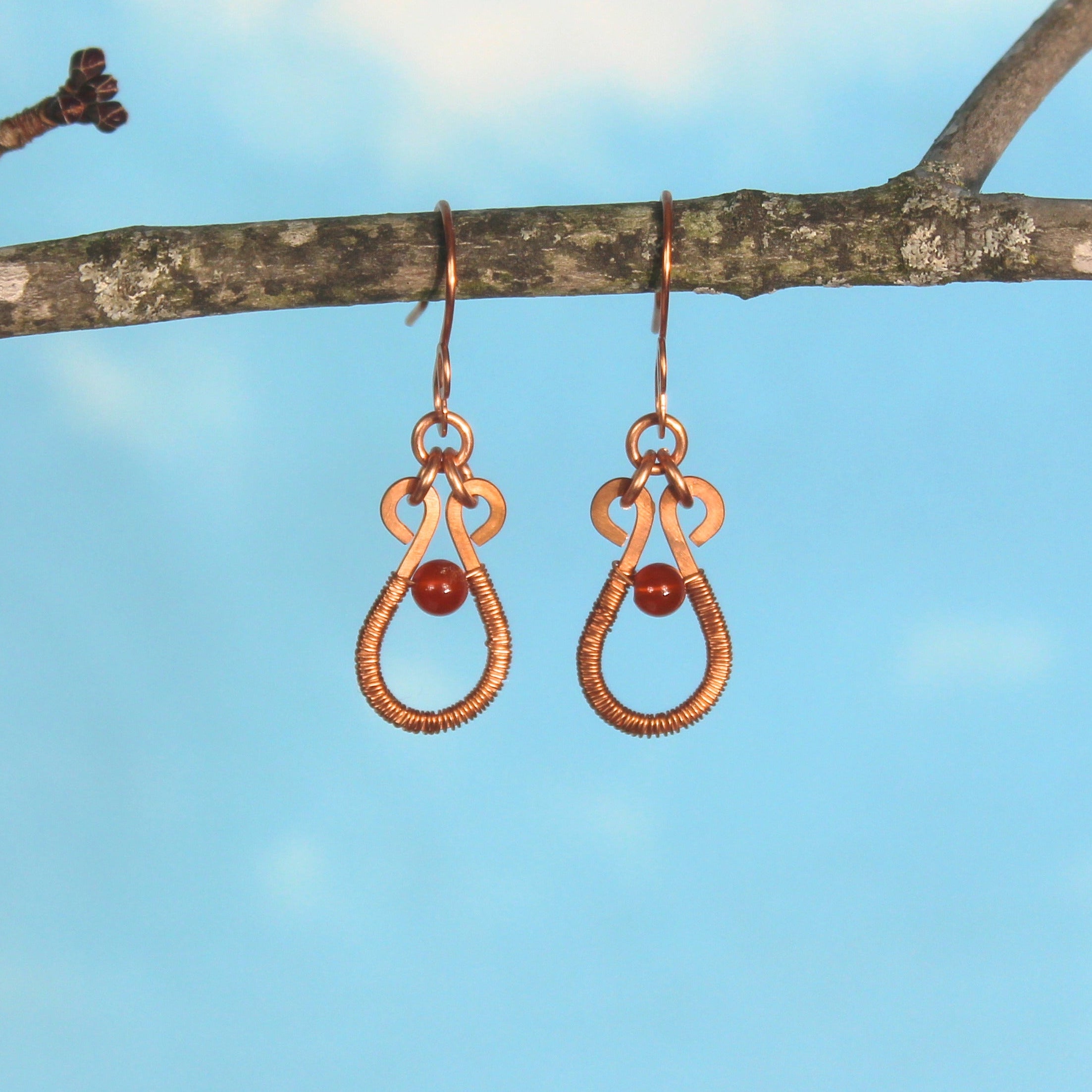 Flat wire copper teardrop earrings wire wrapped with red carnelian bead accents hanging from a branch against a blue sky.