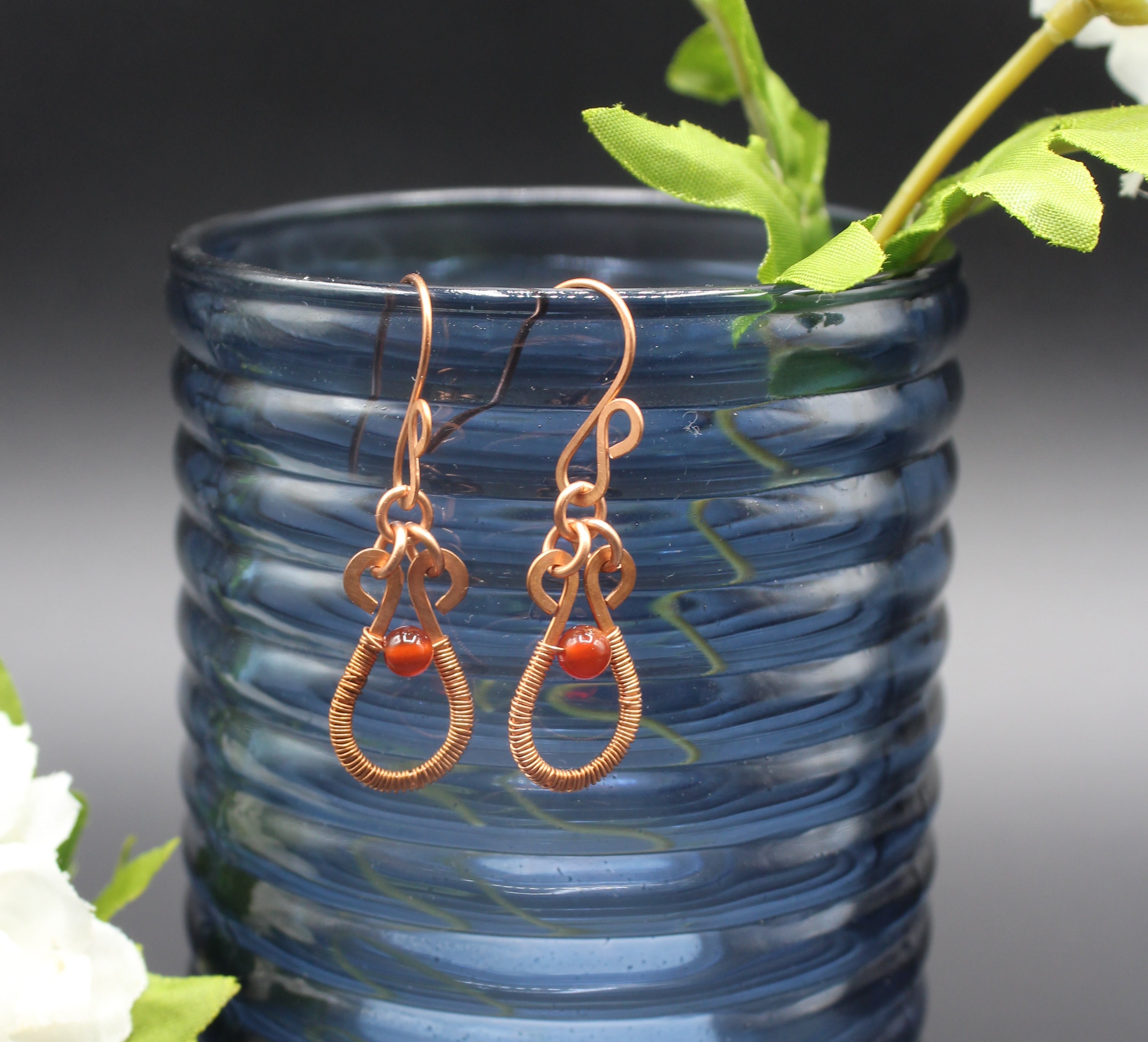 Flat copper teardrop earrings wire-wrapped with red carnelian bead accents in front of a blue glass vase.