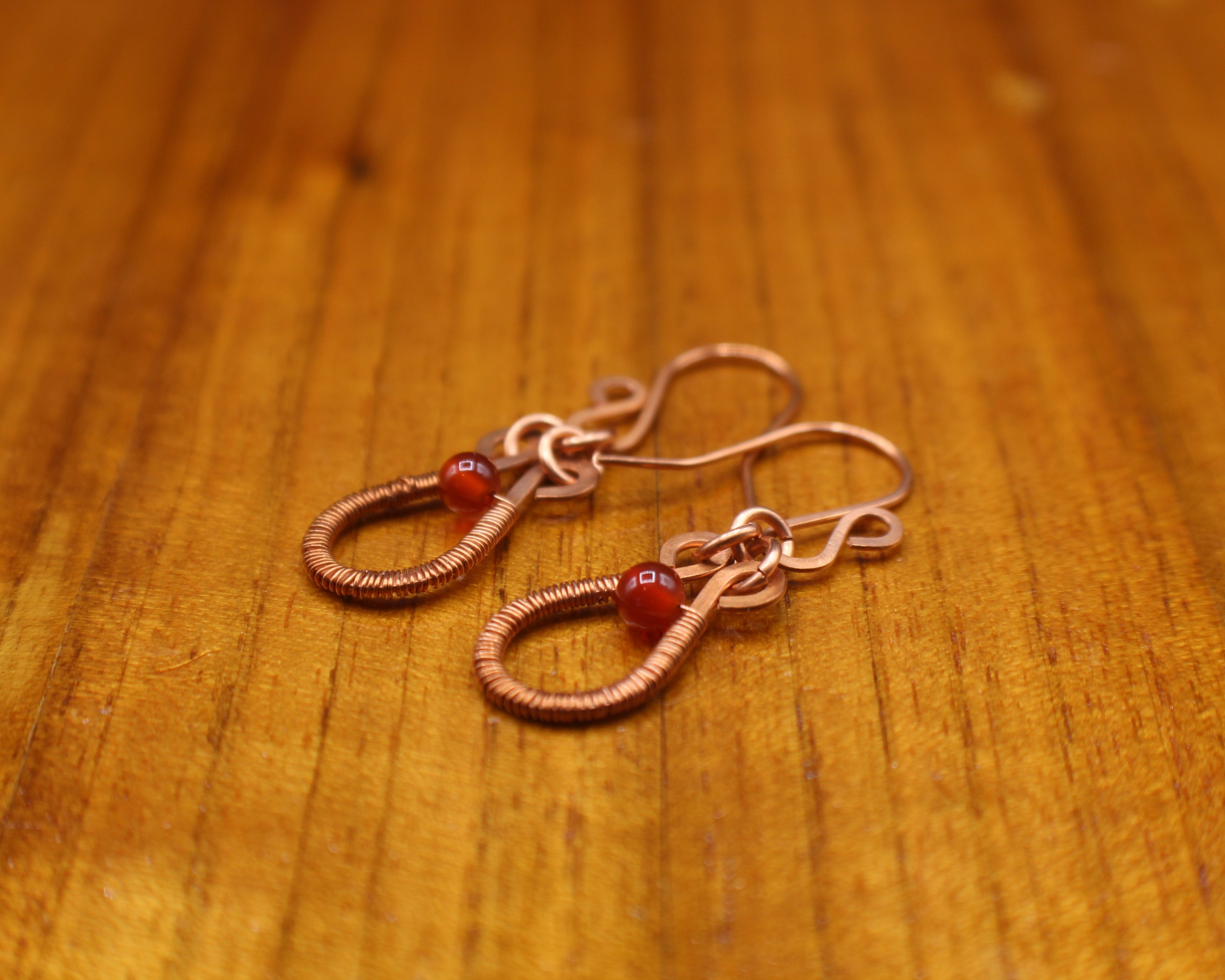 Flat copper teardrop earrings wire-wrapped with red carnelian bead accents  on a wooden surface