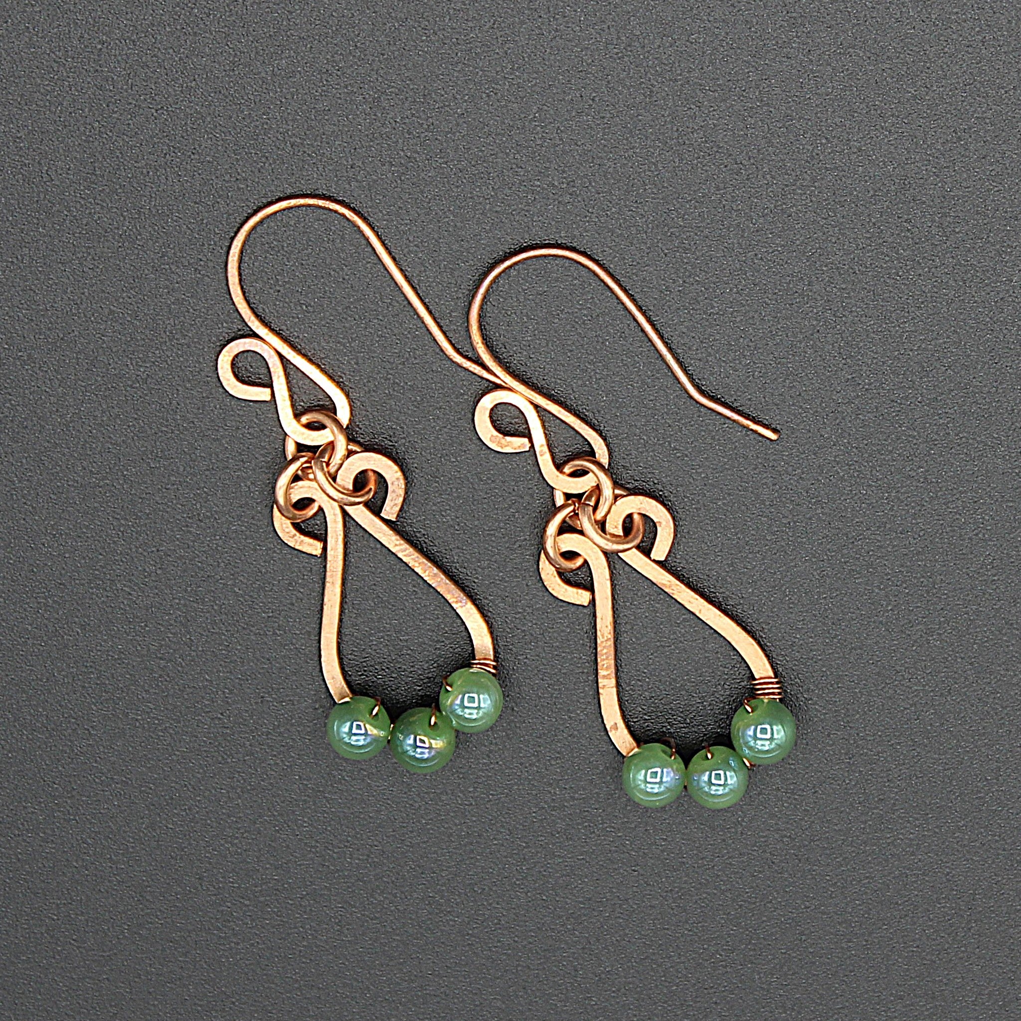 Hammered copper teardrop earrings with small green bead accents on a gray background.