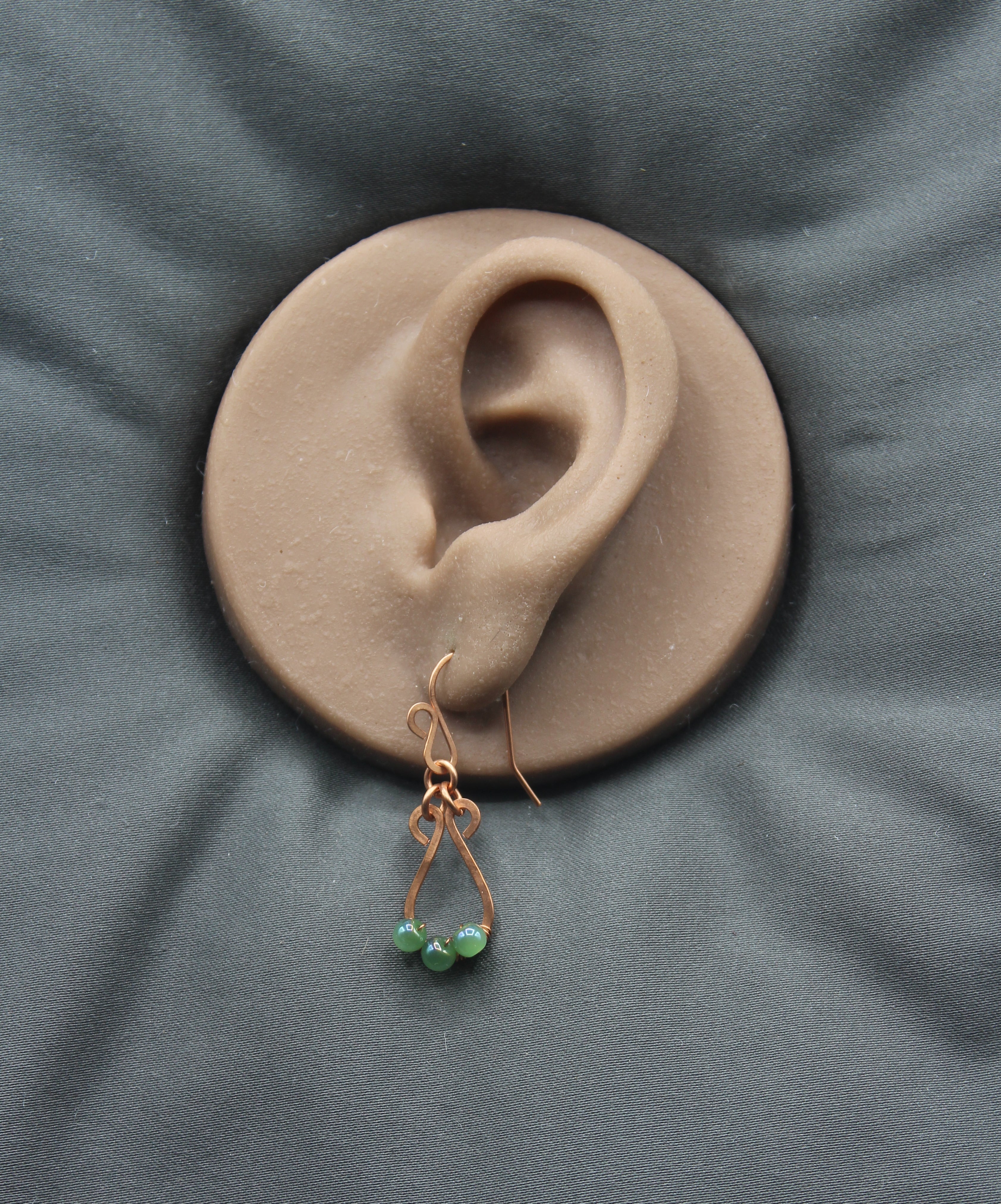 Hammered copper teardrop earrings with small green bead accents on a clay model of an ear against a gray background